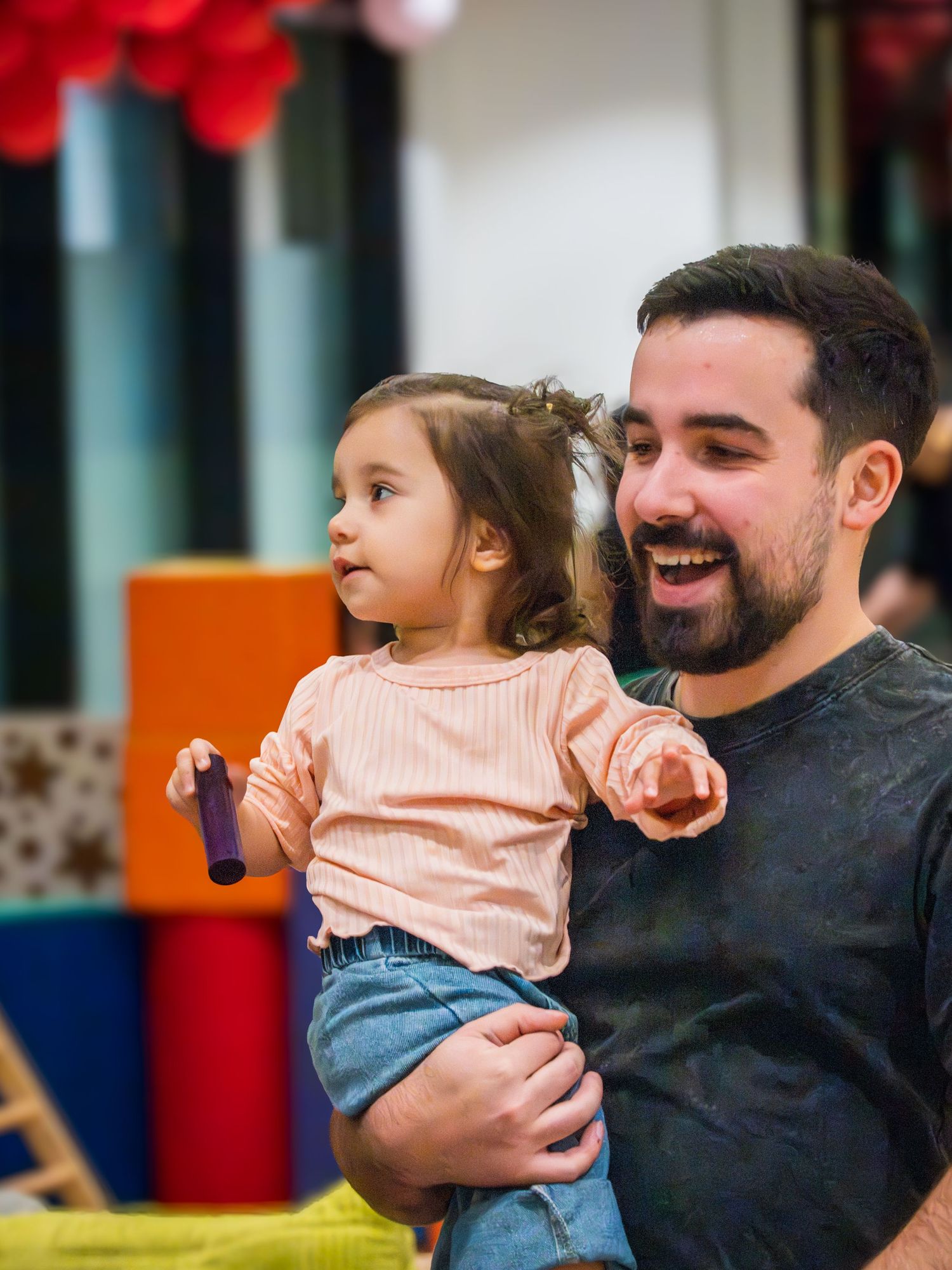 Smiling bearded father holding his young toddler daughter at a colorful indoor birthday party celebration.