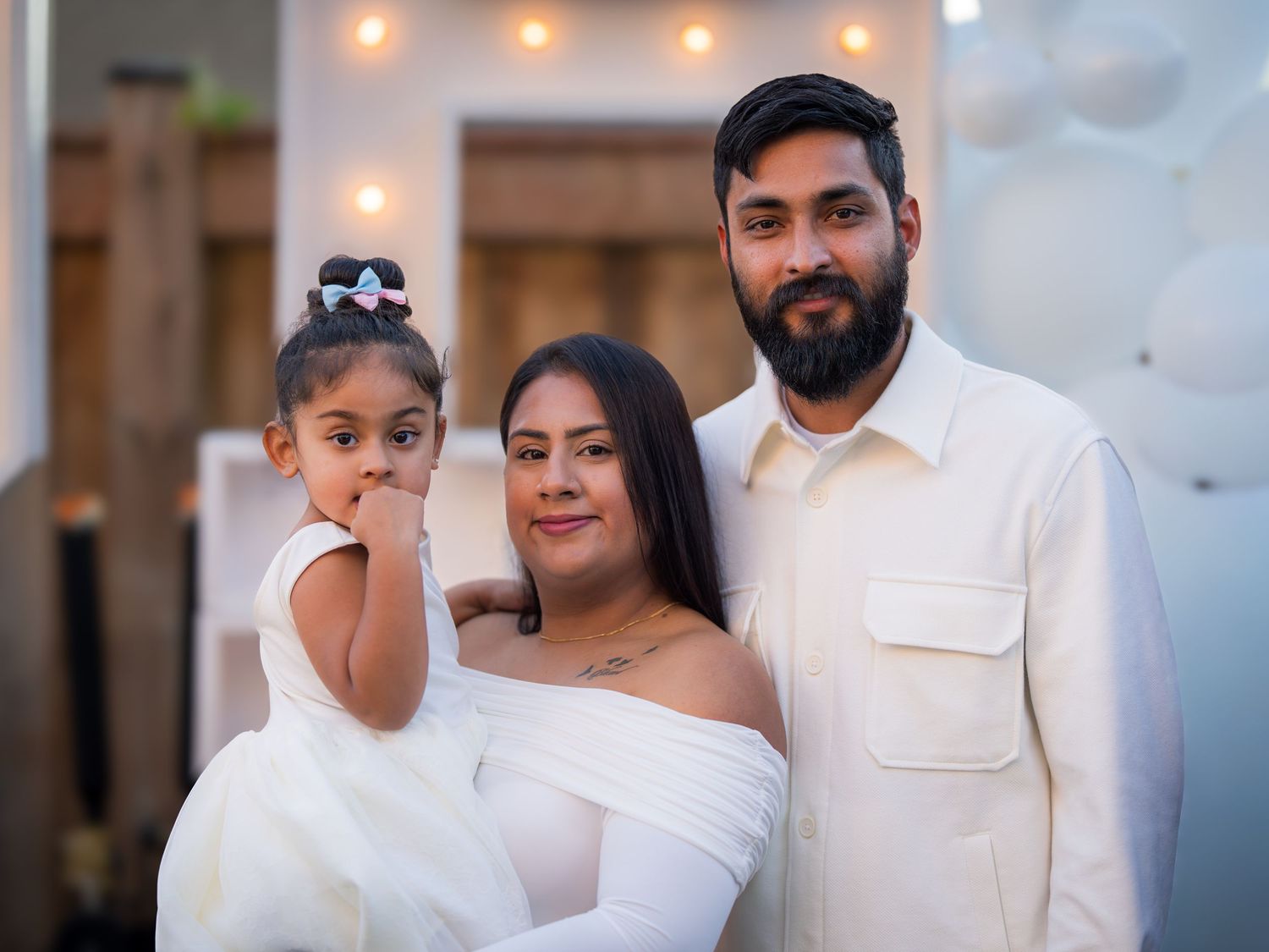 A couple in white outfits poses with their young daughter at a gender reveal celebration.