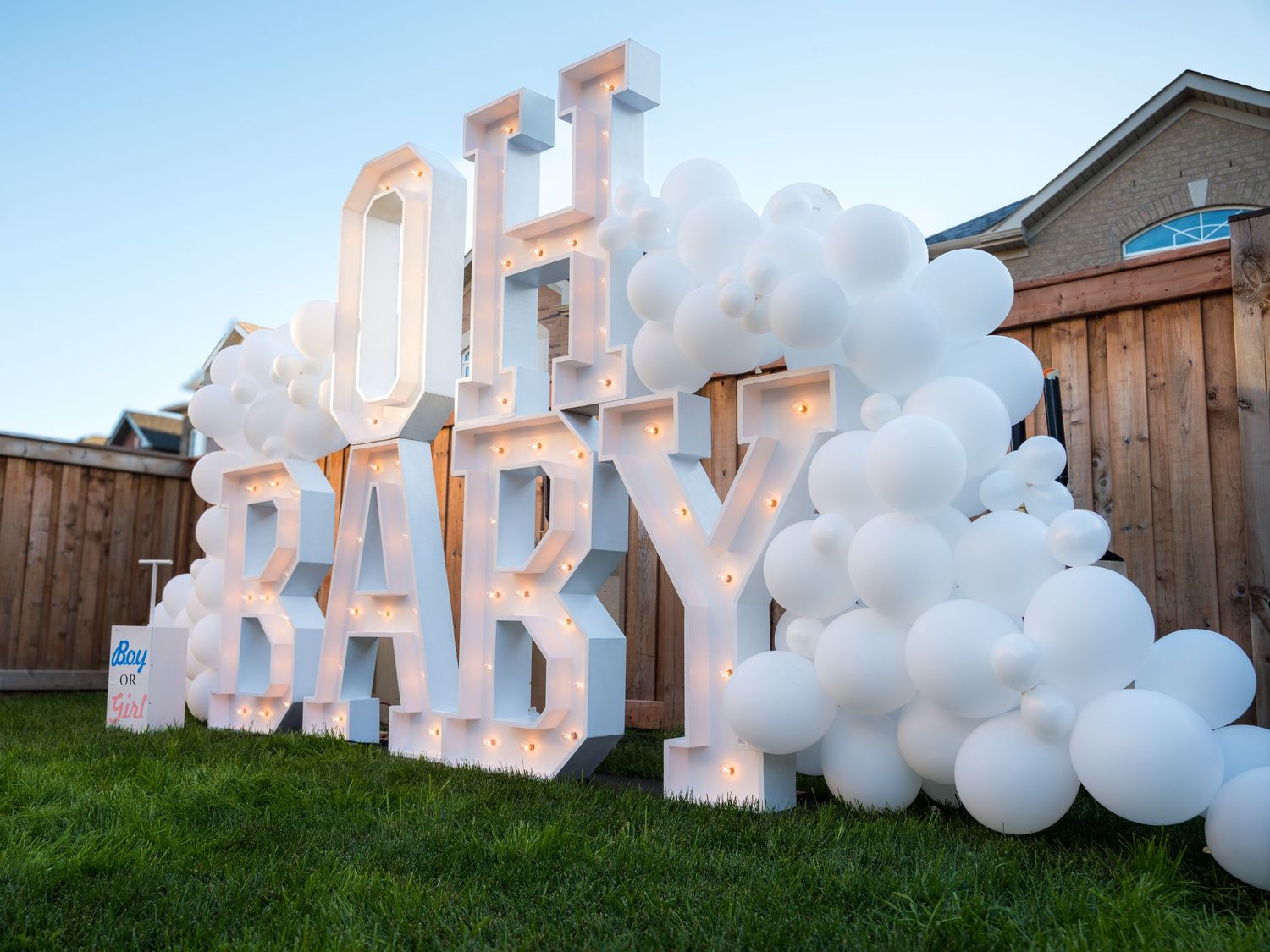 Illuminated OH BABY marquee letter sign surrounded by white balloon arch in backyard at dusk.