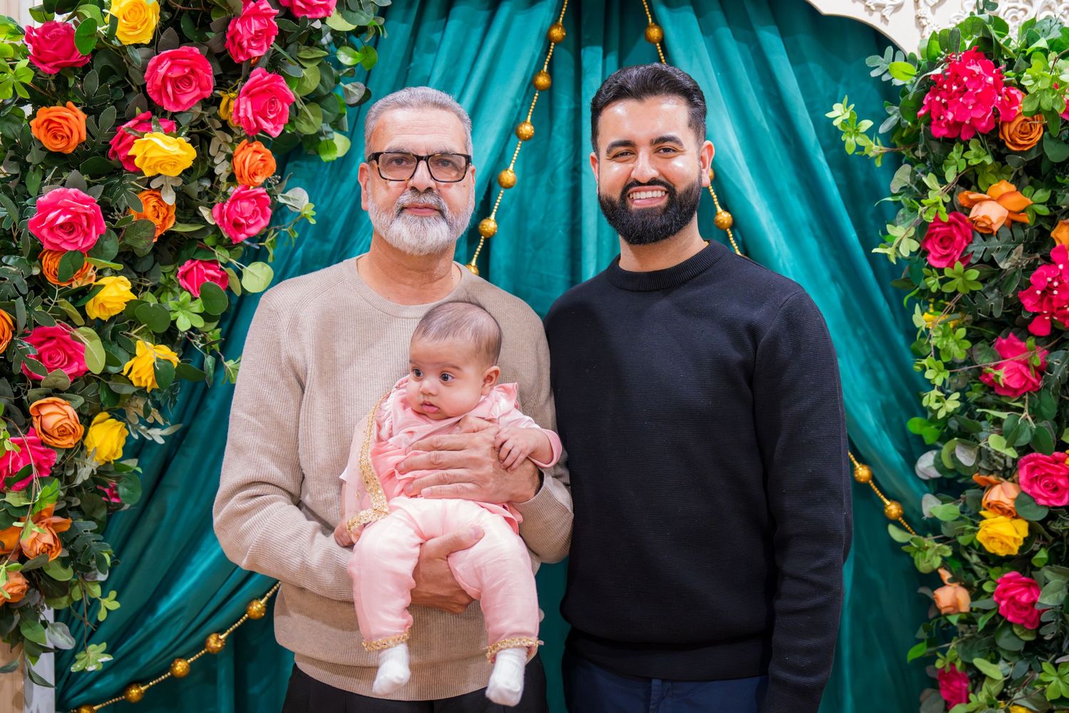 Two men, one holding a newborn baby in pink, pose in front of a colorful floral arch with teal draping.