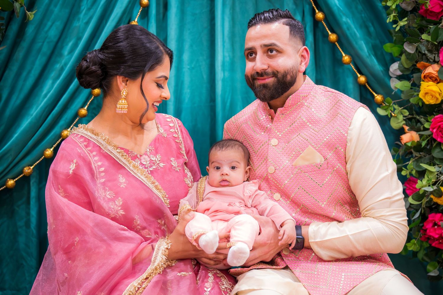 South Asian parents in traditional pink and cream attire lovingly holding their infant at a festive event.