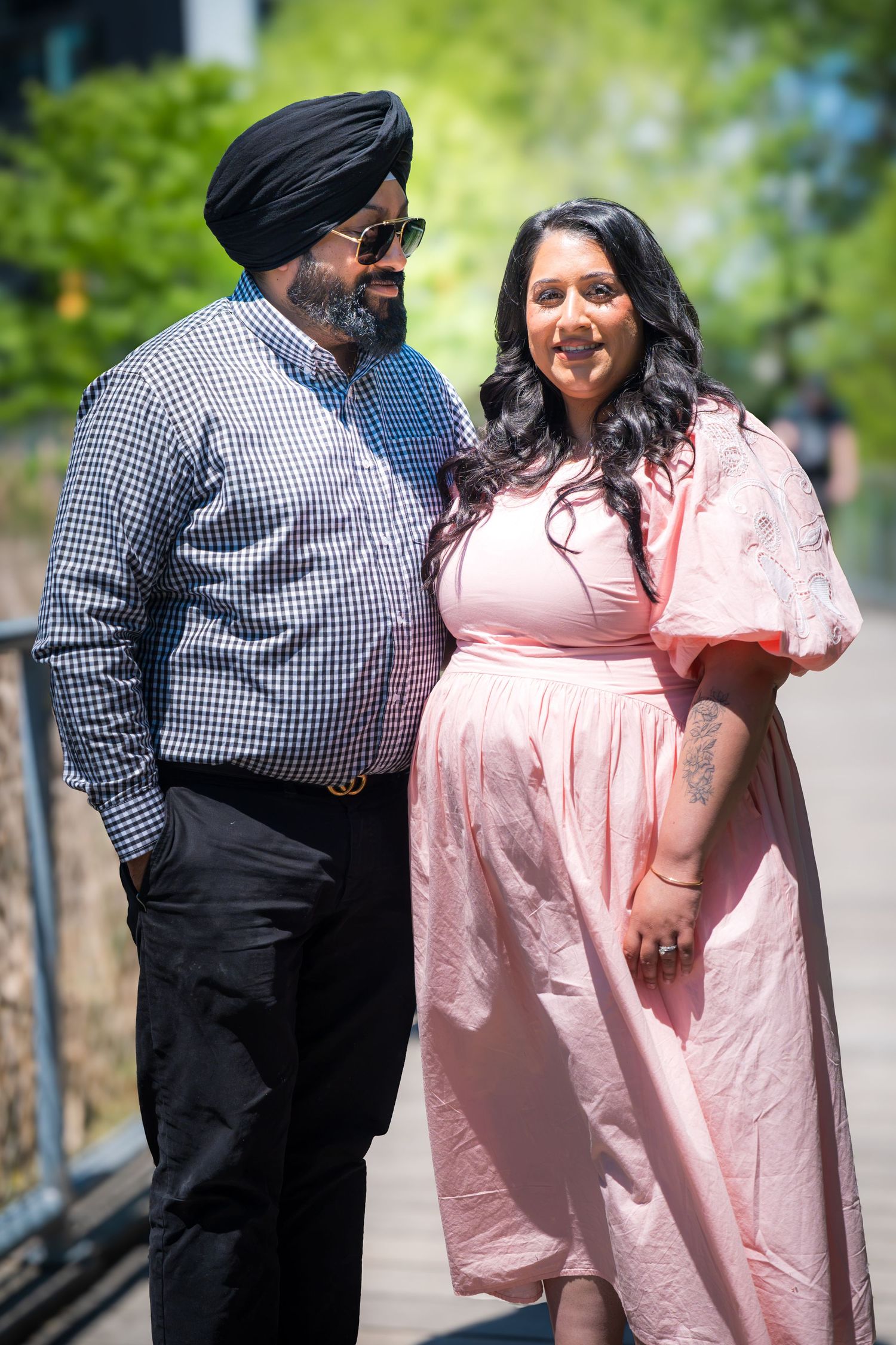 A Sikh man in a turban and sunglasses stands with a woman in a pink dress on a sunny urban walkway.