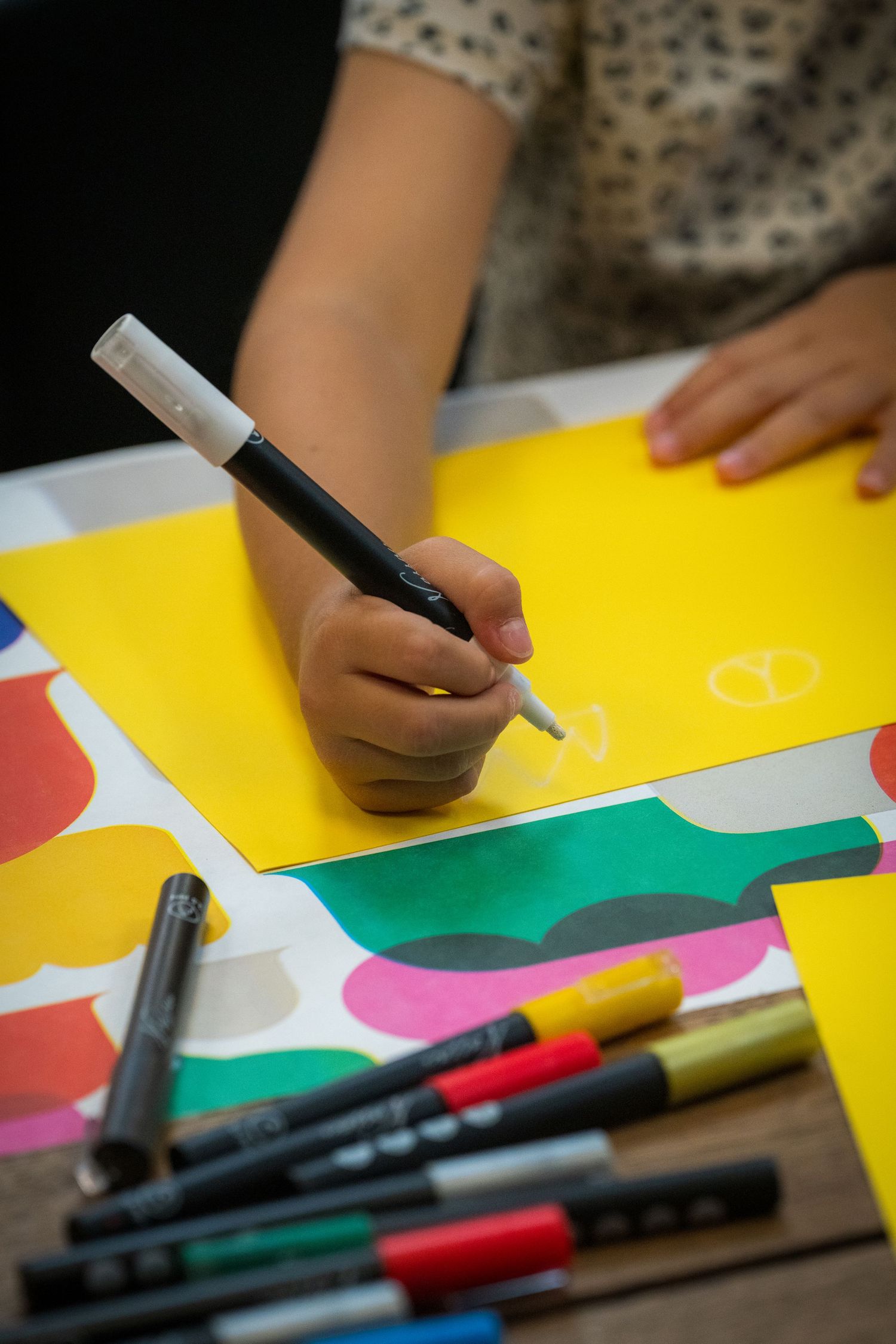 Child's hand drawing with white marker on yellow paper surrounded by colorful markers and paper shapes.