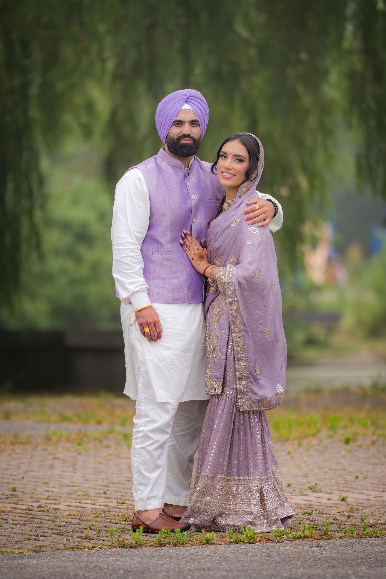 Couple in traditional Punjabi dress poses lovingly under willow trees during outdoor photoshoot.