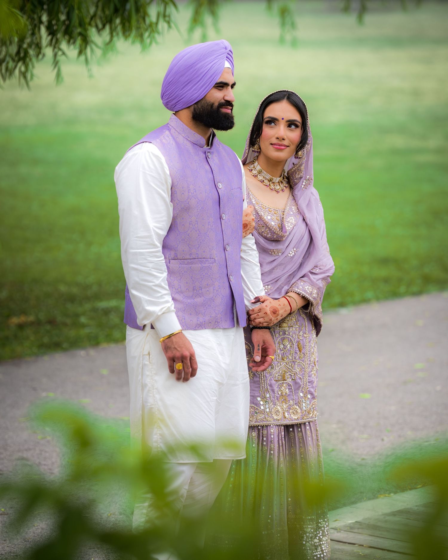 A couple in traditional Punjabi dress standing on a path with greenery in the foreground.