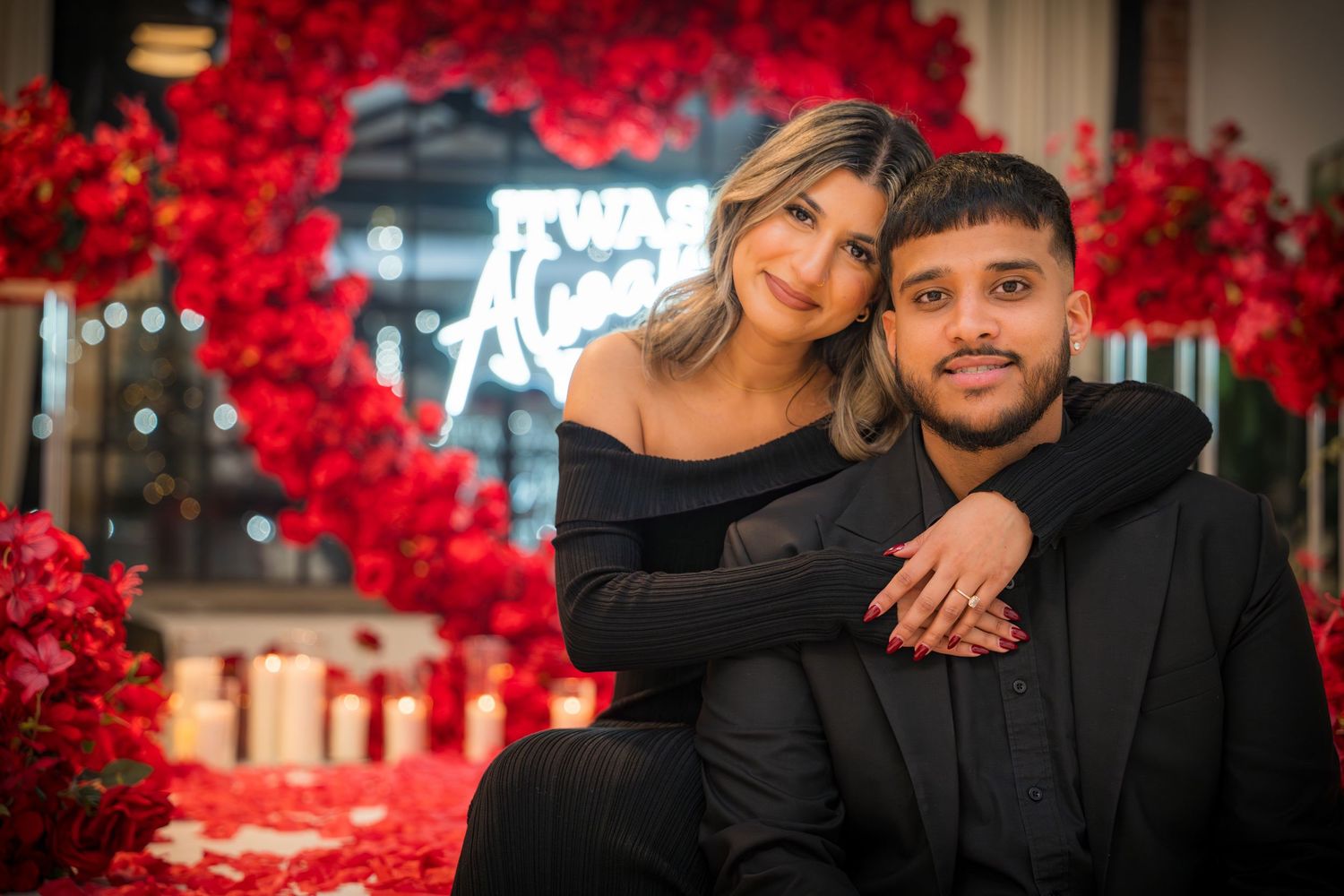 Couple embracing in front of romantic red rose wall with neon lighting.
