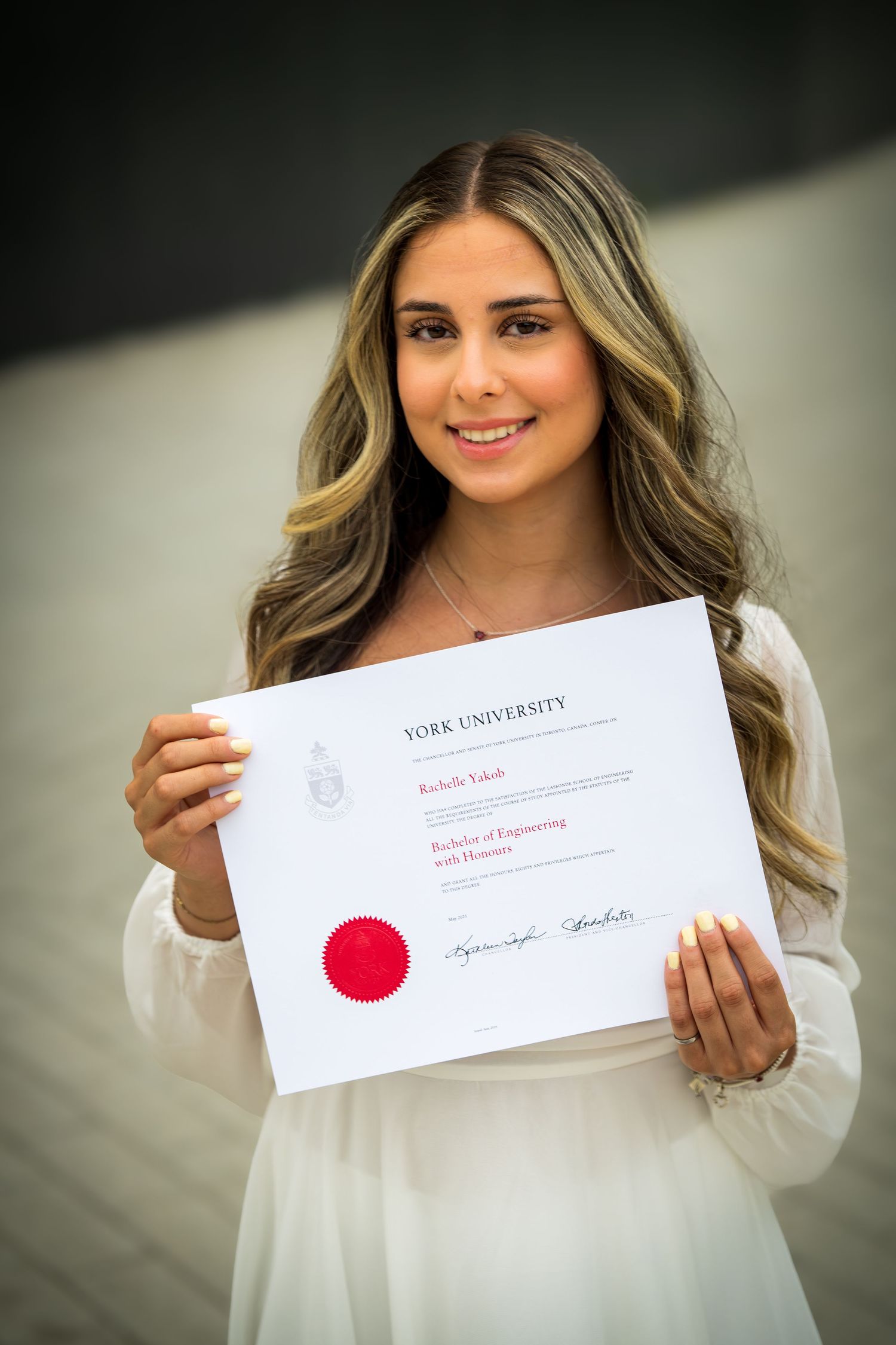 York University graduate holding diploma certificate while wearing elegant white dress.