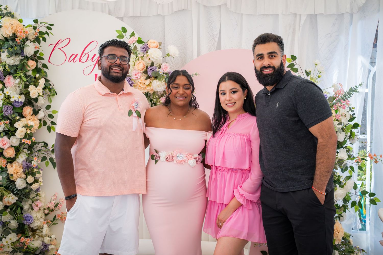 Four guests pose together at baby shower with floral arch and white draping.