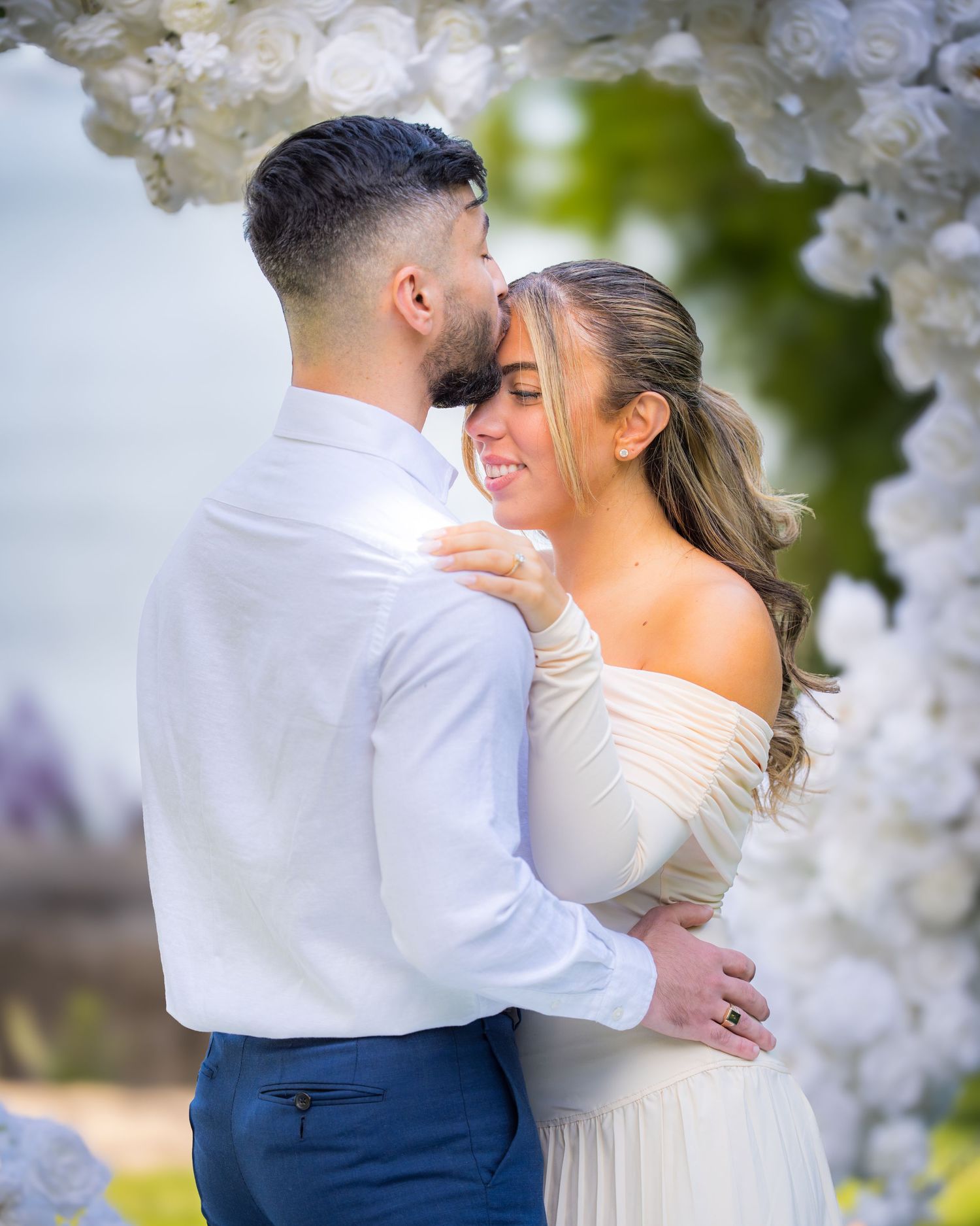 Couple embracing under an elegant white floral arch at an outdoor wedding venue.