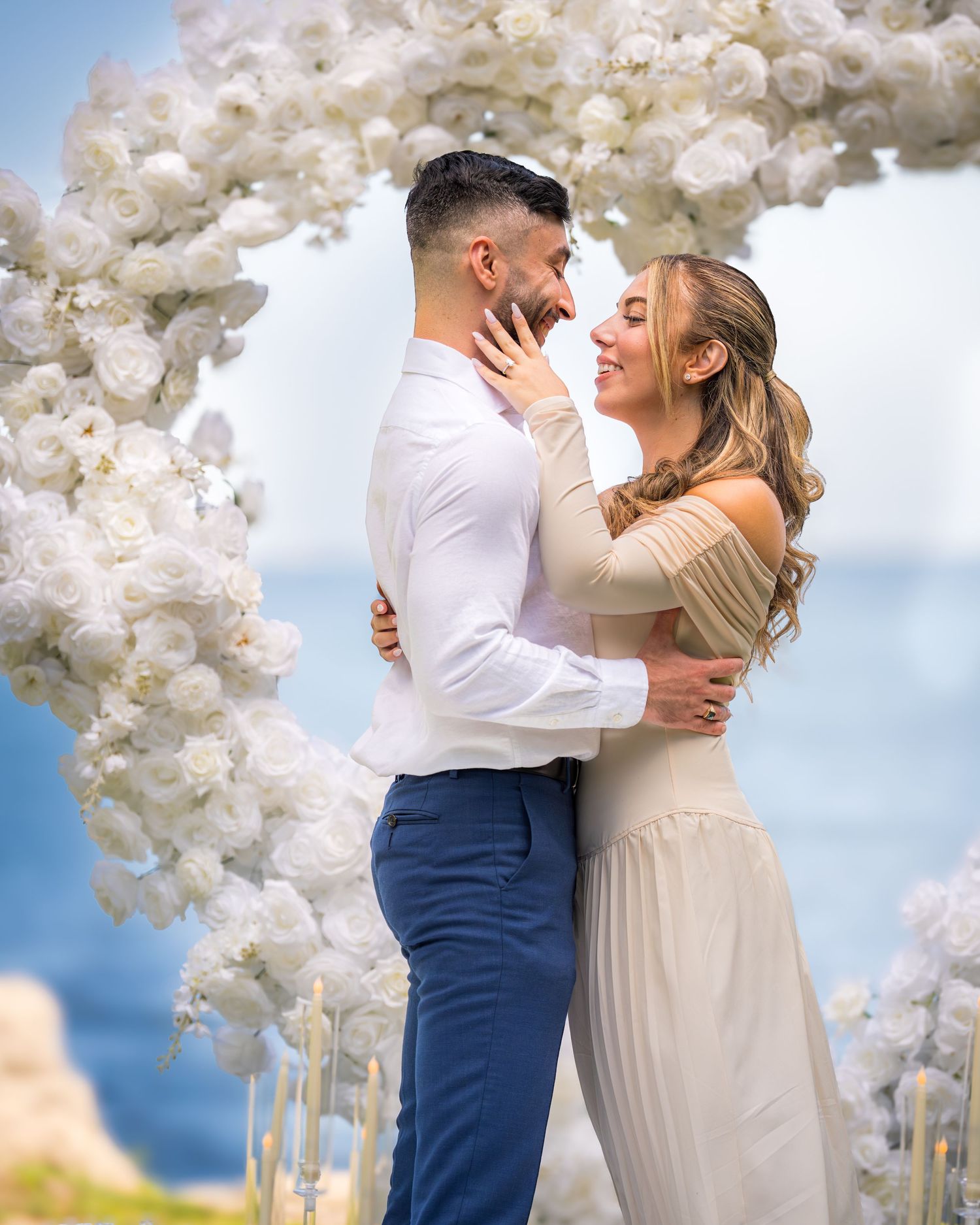 Romantic couple embracing under white floral arch with ocean view in background.