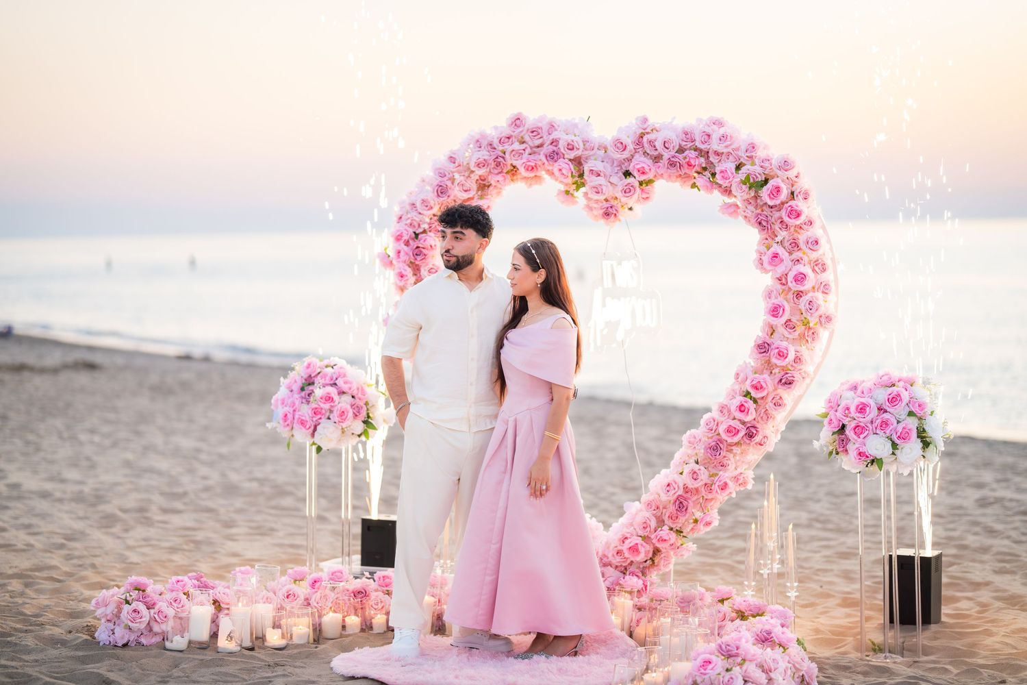 Pink floral heart arch and decorative elements arranged on beach at sunset.