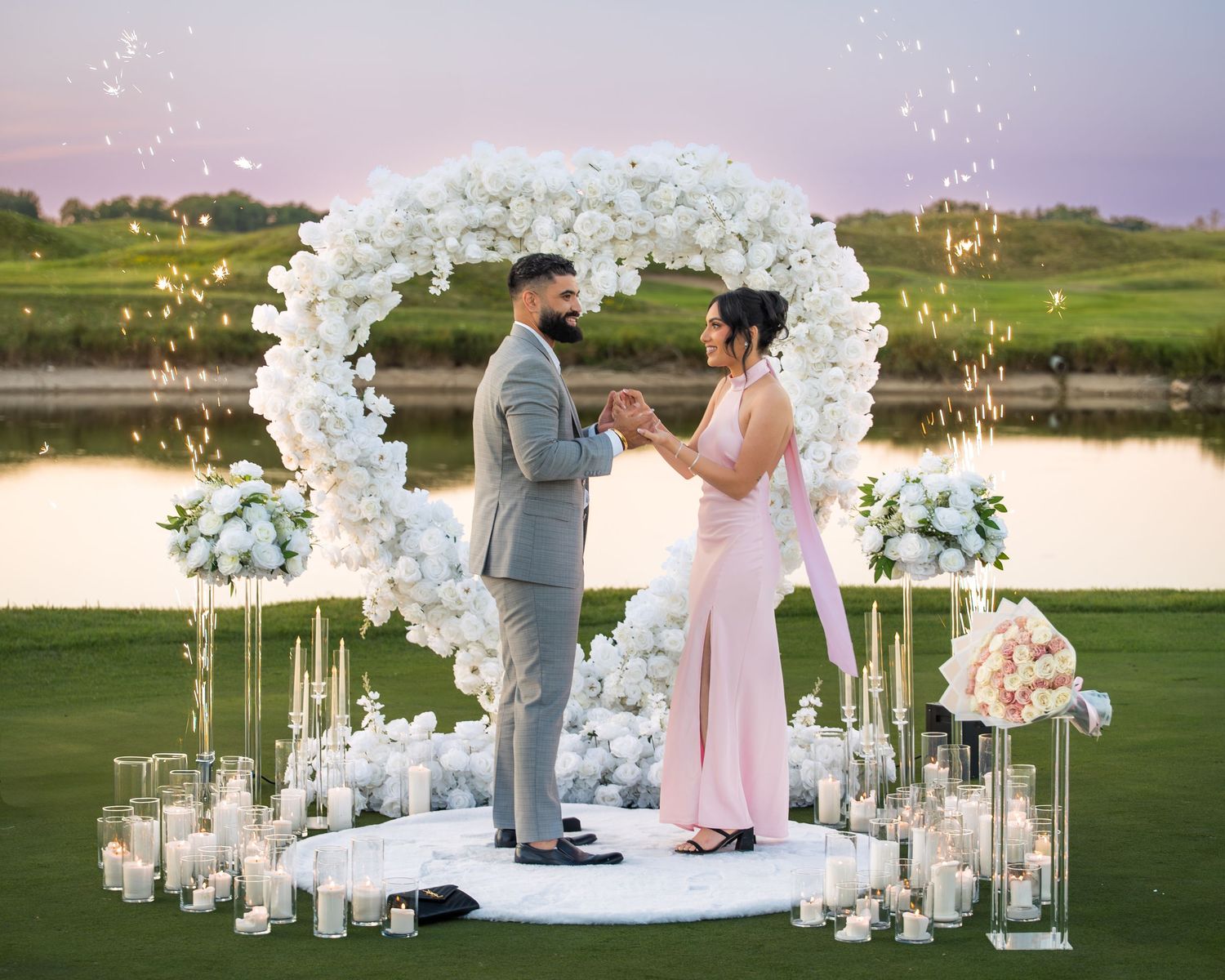 Romantic outdoor proposal scene with white floral decorations and candlelit pathway at sunset.