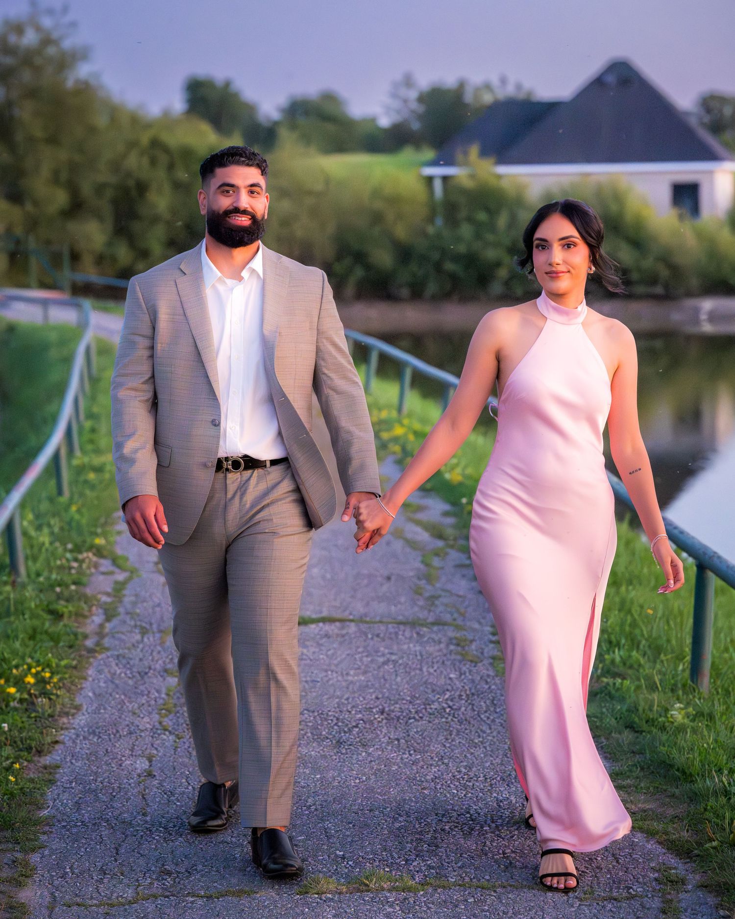 A couple walks hand in hand along a lakeside path, one in a tan suit and one in a pink dress.