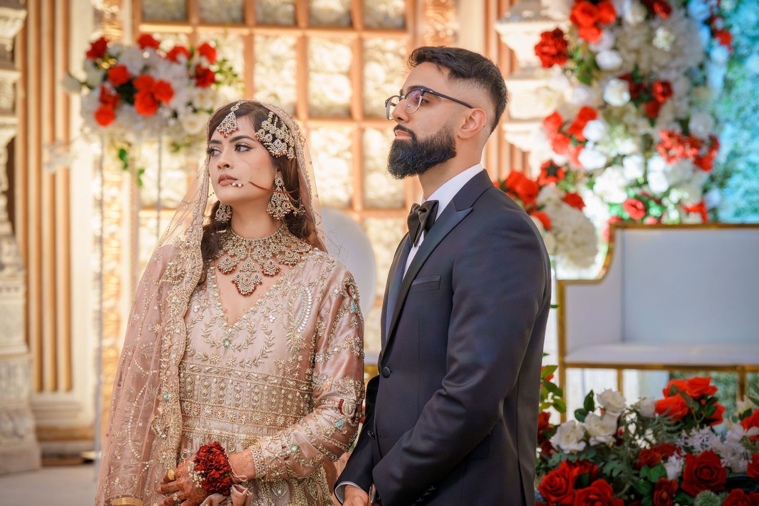 Wedding couple in traditional South Asian attire pose against colorful floral backdrop.