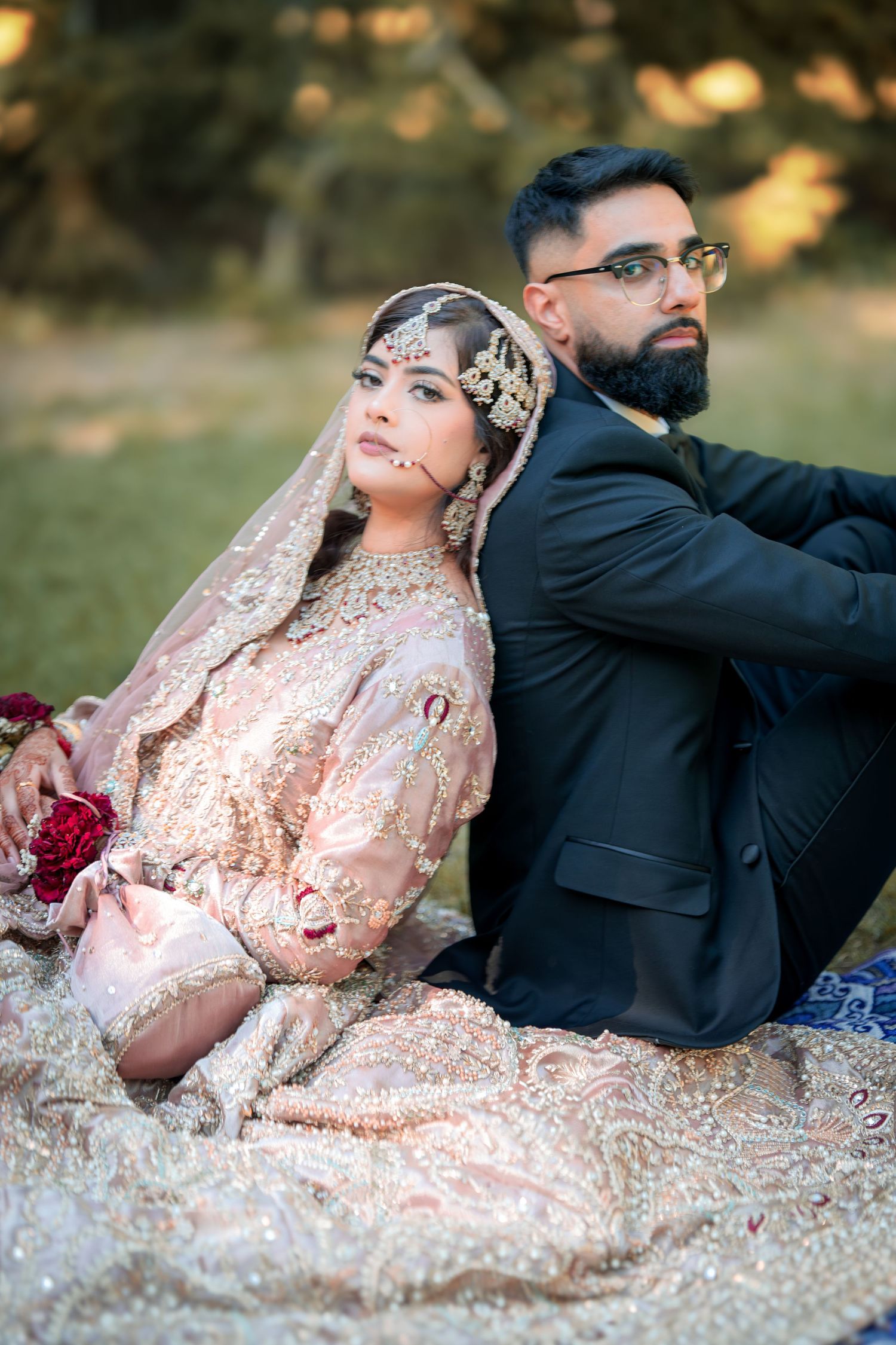 A South Asian couple in wedding attire poses outdoors with bride in ornate pink lehenga and groom in black suit.