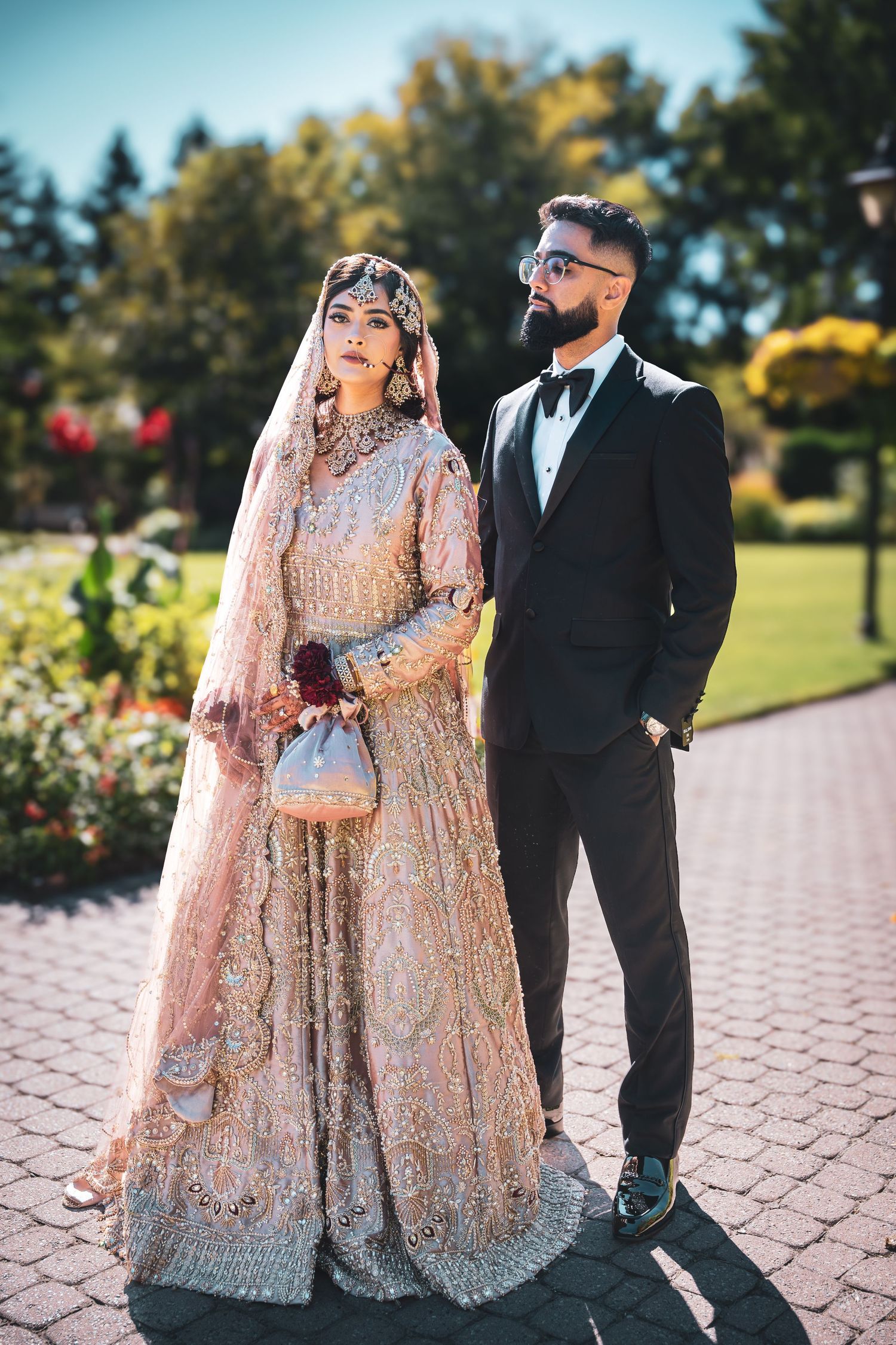 A couple in traditional South Asian wedding attire stands on a paved walkway.