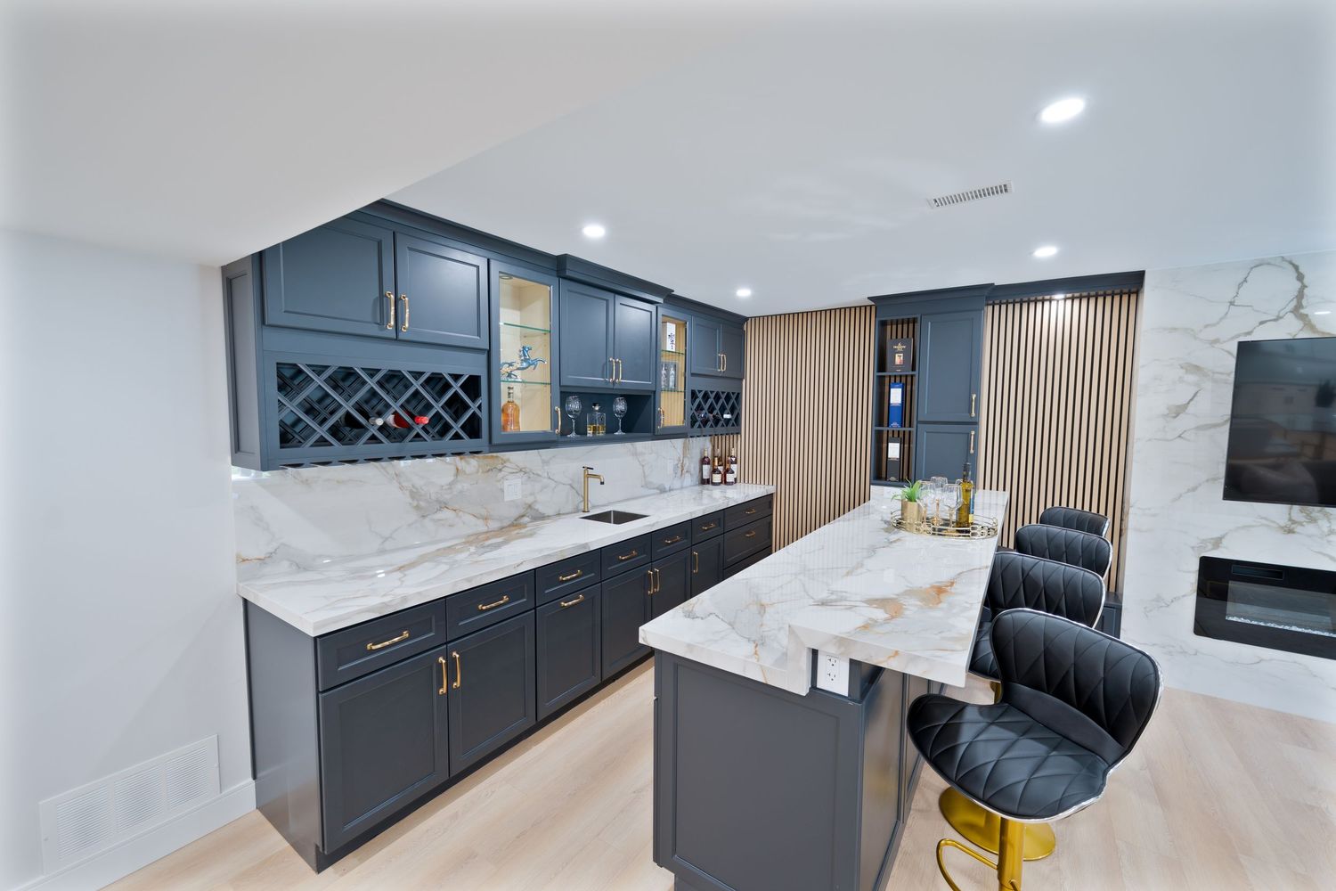 Modern basement bar features dark gray cabinets, marble countertops, wine storage, and velvet bar stools in a luxurious setting.