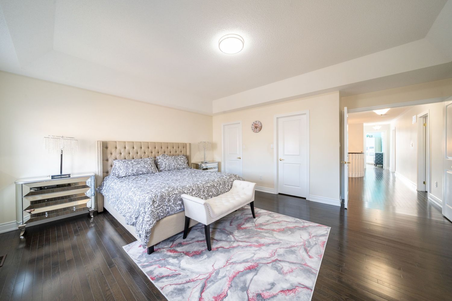 Modern bedroom interior with tufted headboard, mirrored nightstand, and pink area rug on dark hardwood floors.