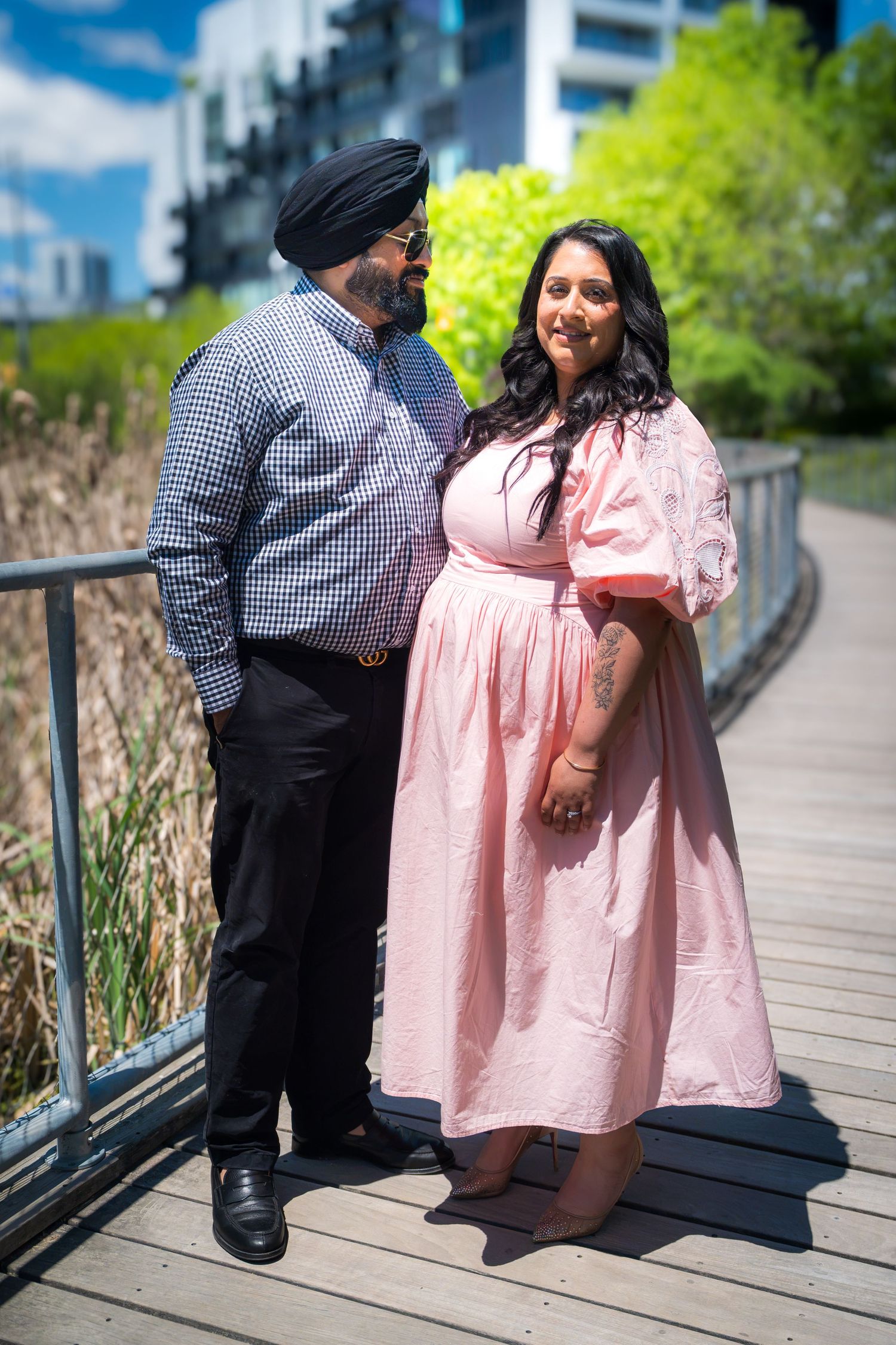 Couple posing on wooden bridge, man in turban and plaid shirt, woman in pink dress, urban park setting.
