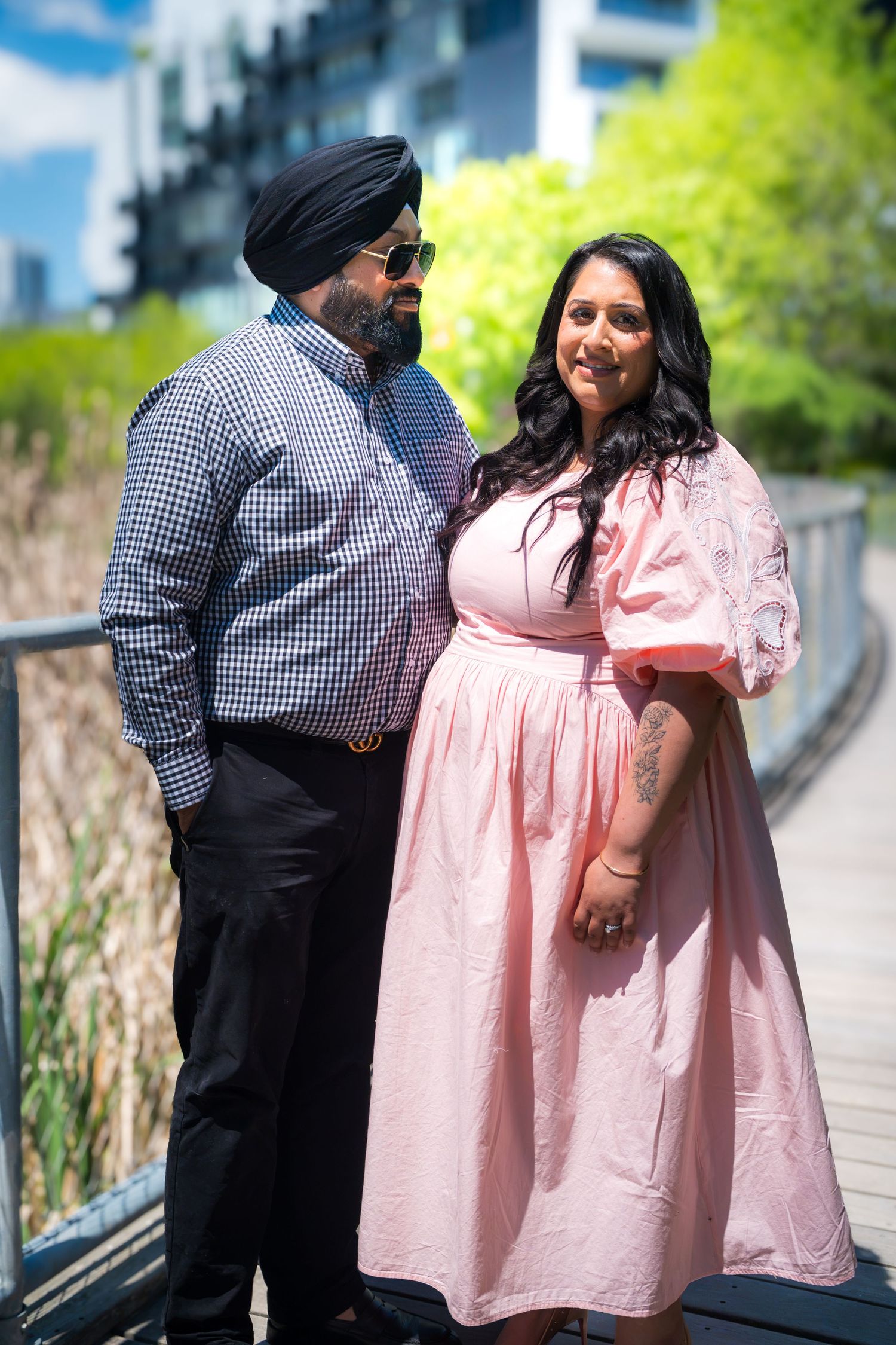 Sikh man in turban and woman in pink maternity dress posing together on a bridge outdoors.