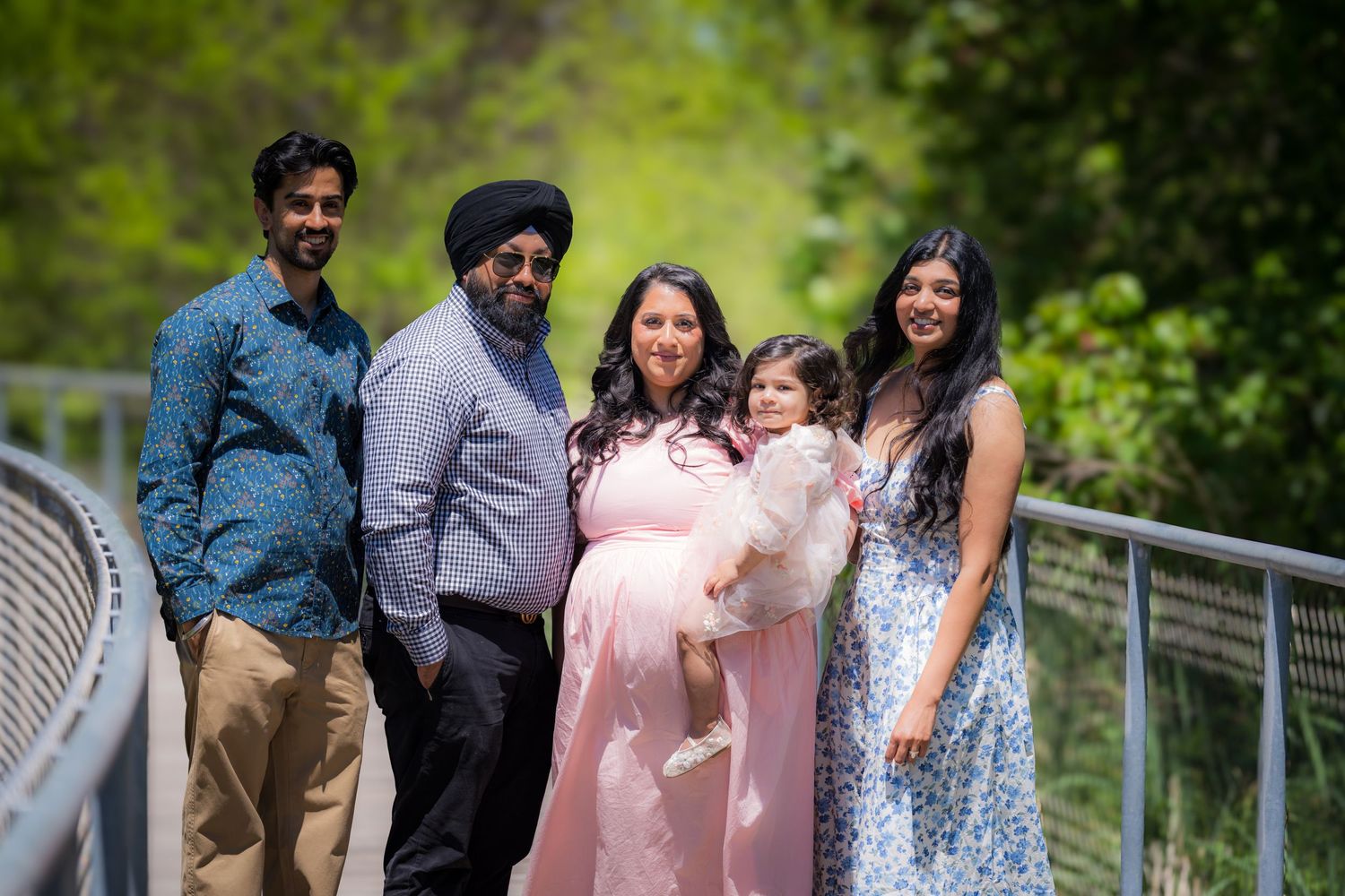 Extended Sikh family of five posing together on a bridge surrounded by green trees outdoors.