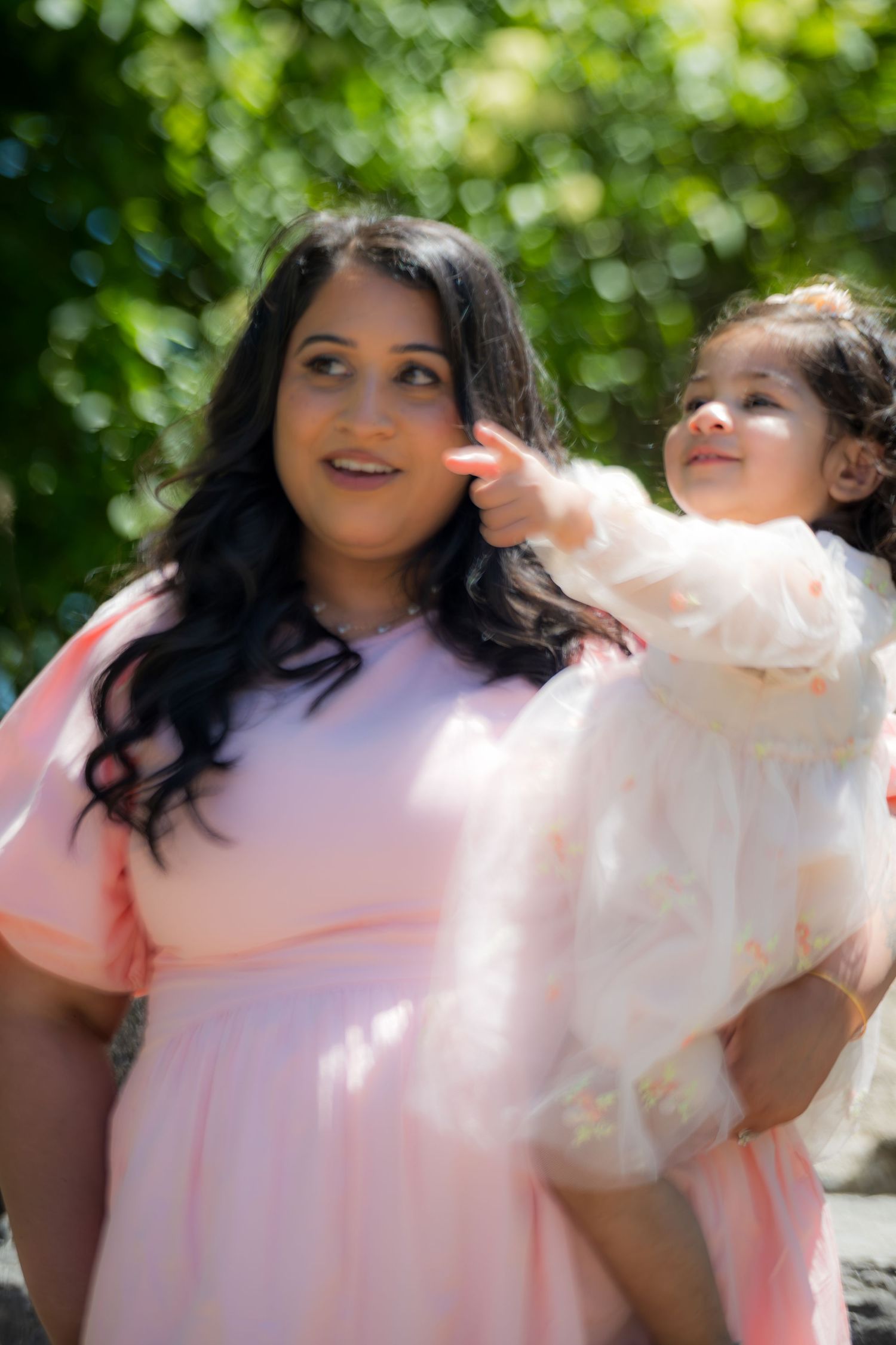 Woman in pink dress holds toddler in white outfit outdoors in lush green park setting.