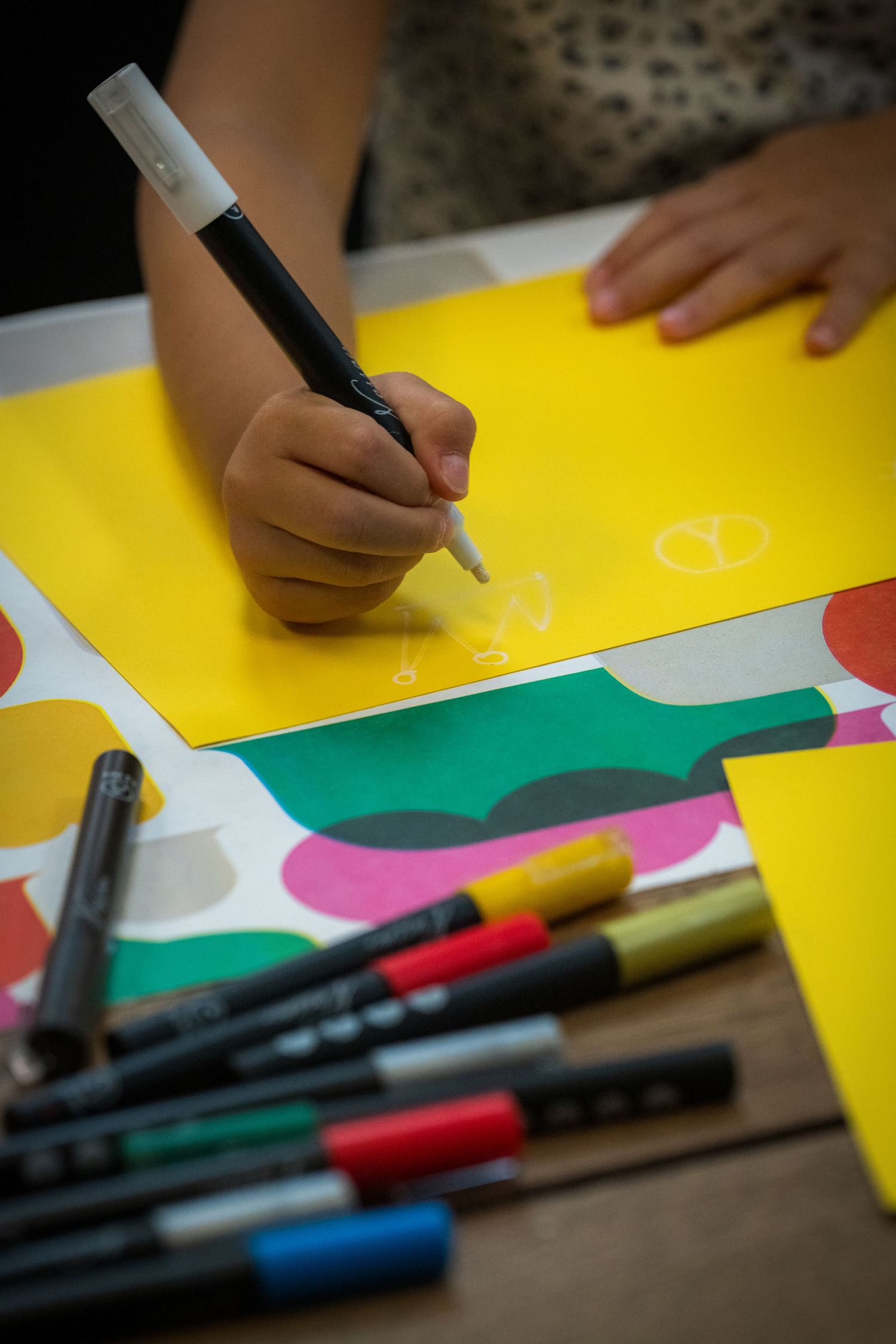 Child draws on yellow paper with marker surrounded by colorful art supplies.
