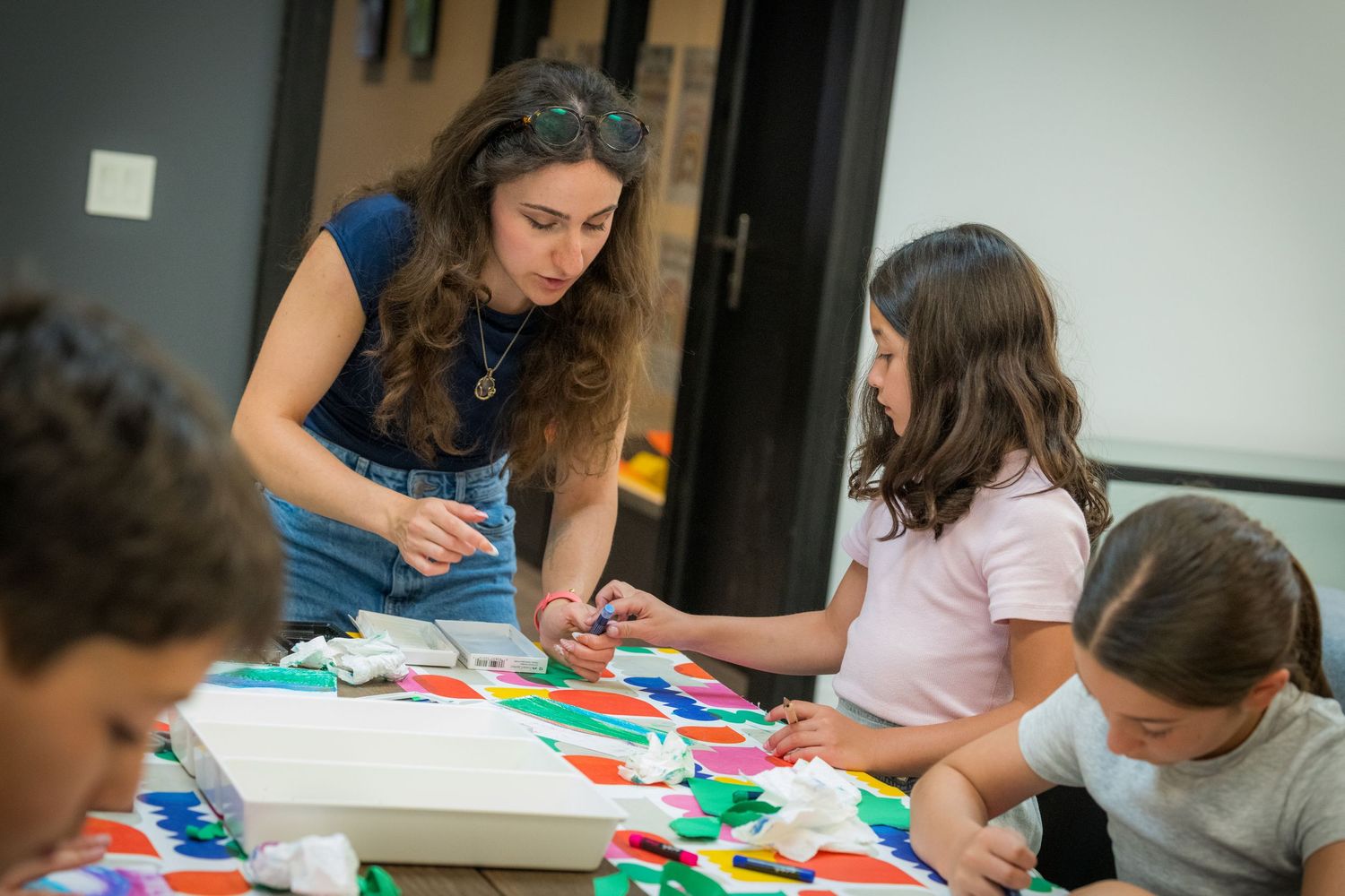 A woman guides two kids in a colorful paper craft project at a bright table.