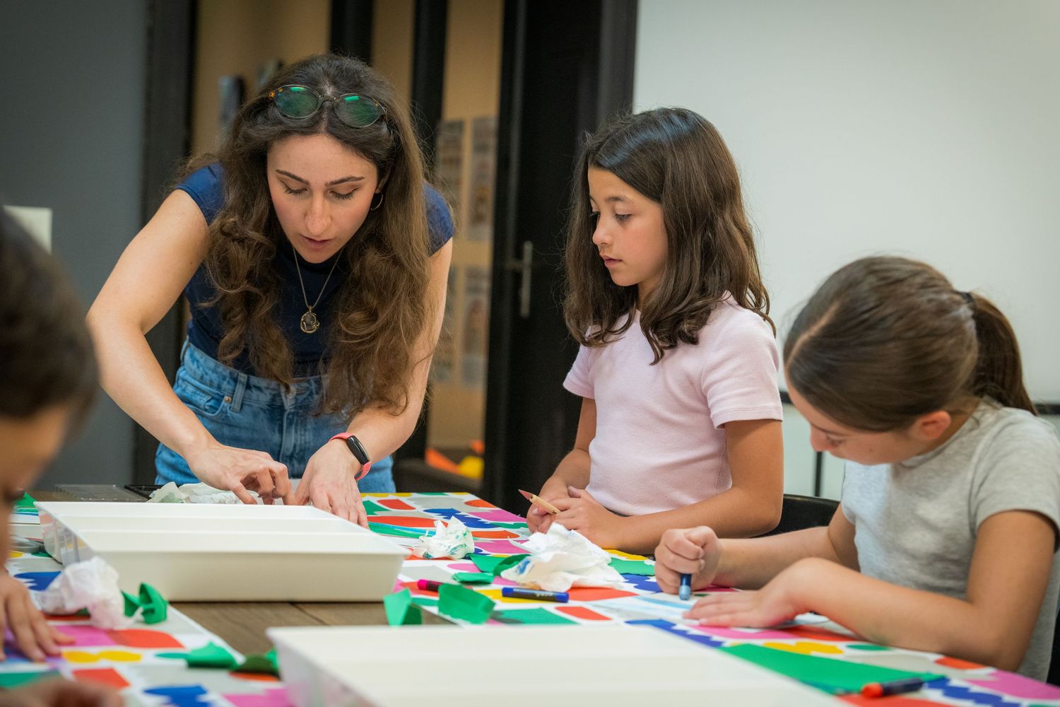 A teacher guides three young girls in a colorful paper craft activity at an art class table.