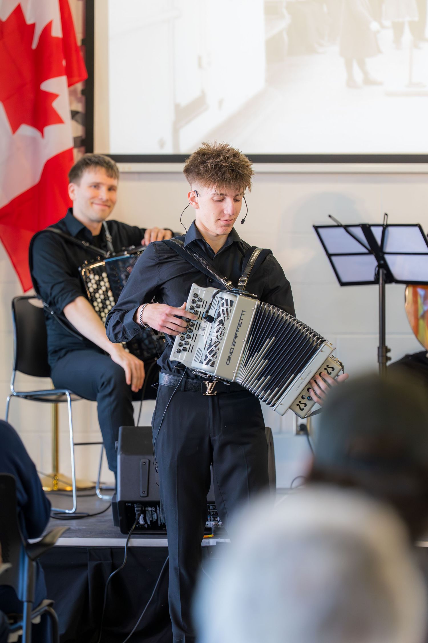 Young male accordionist performing on stage with another accordion player seated behind him.