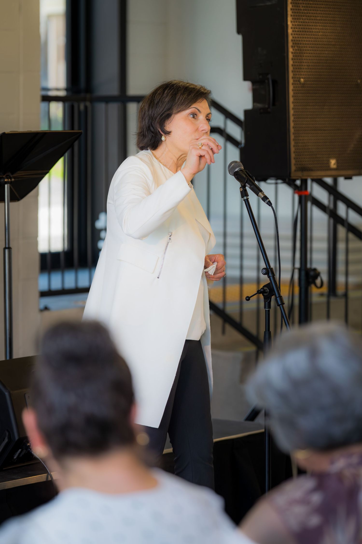 Woman in white blazer speaking at microphone stand during indoor event with audience seated in foreground.