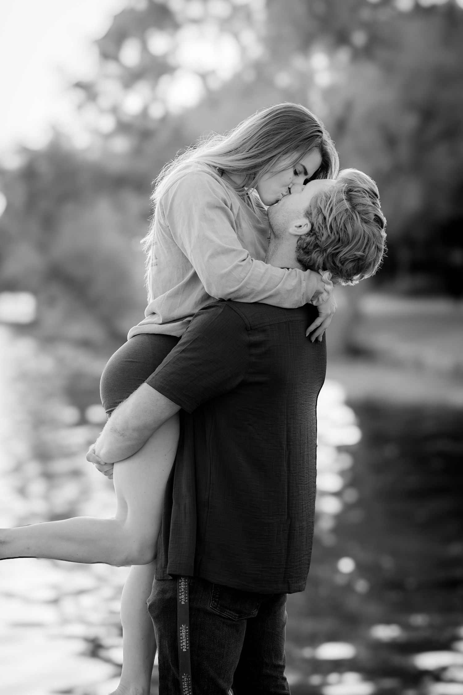 Black and white photo of couple kissing near water, man lifting woman in romantic outdoor setting.
