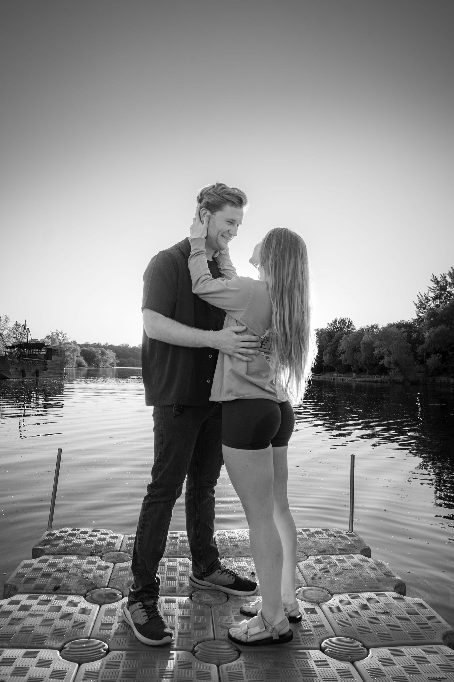 Black and white photo of a couple sharing a tender moment on a dock overlooking a peaceful lake.