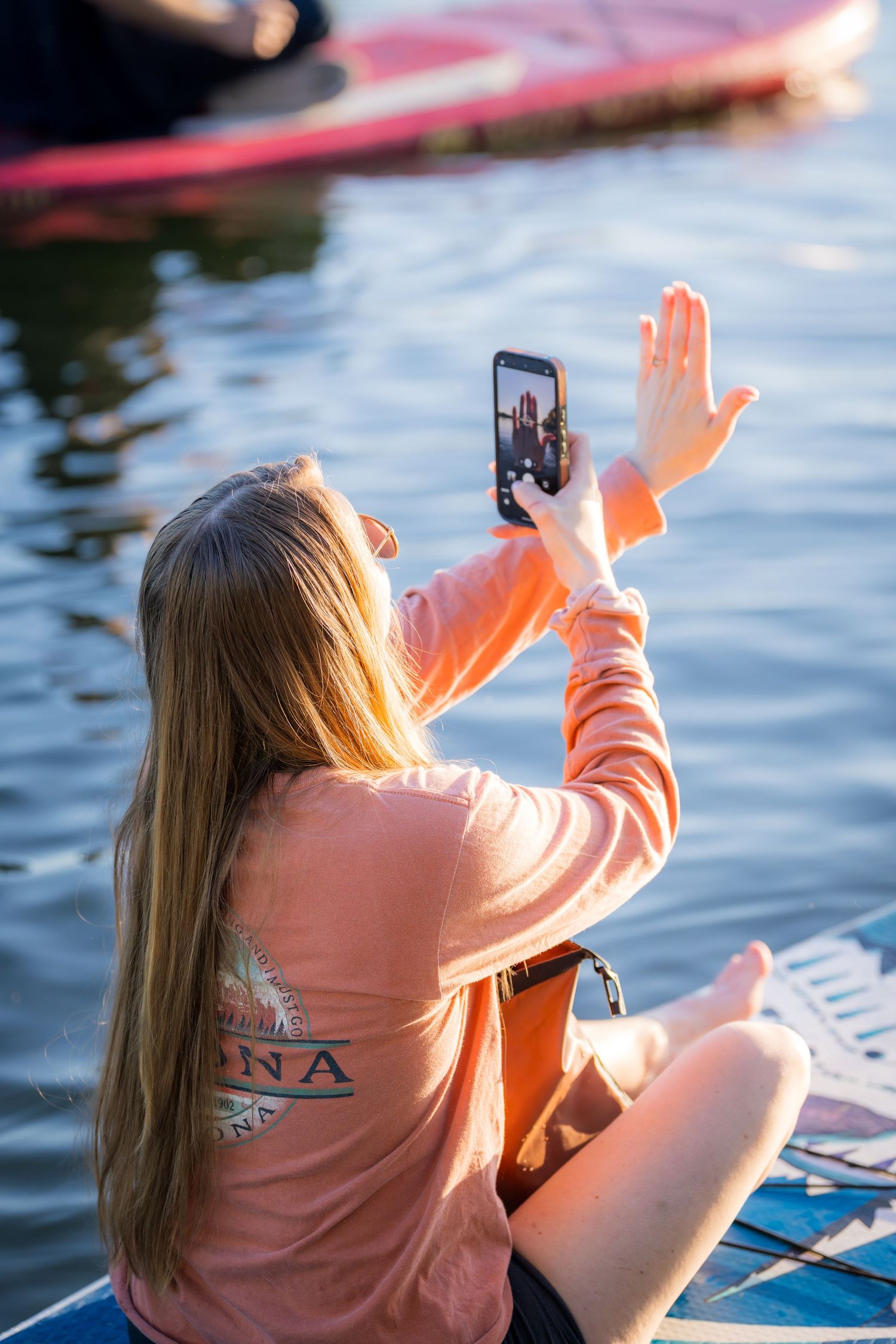 Blonde woman in pink sweatshirt sitting on paddleboard, waving during video call on smartphone near water.
