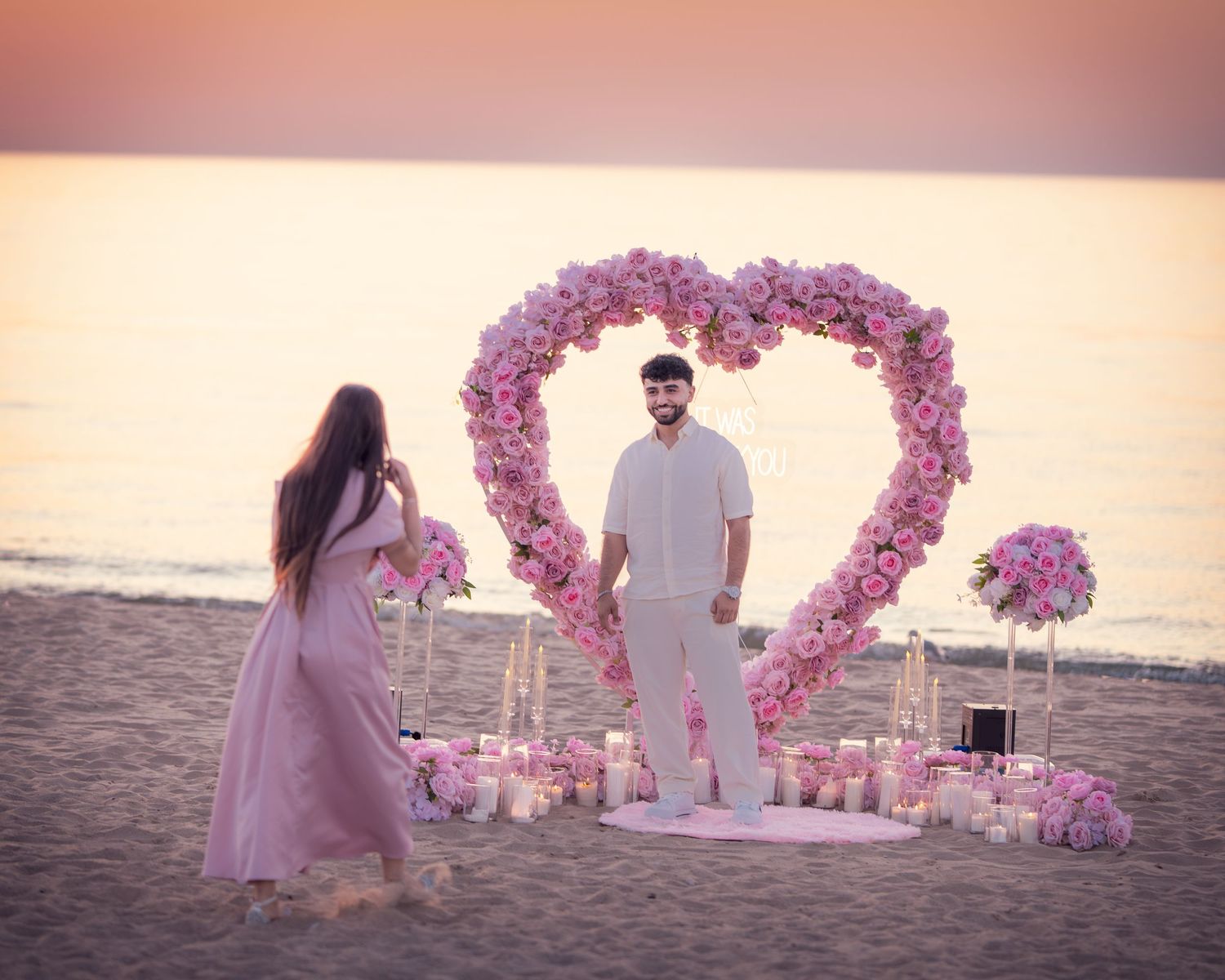 A romantic beach proposal setup with a pink floral arch and couple silhouetted against the sunset.