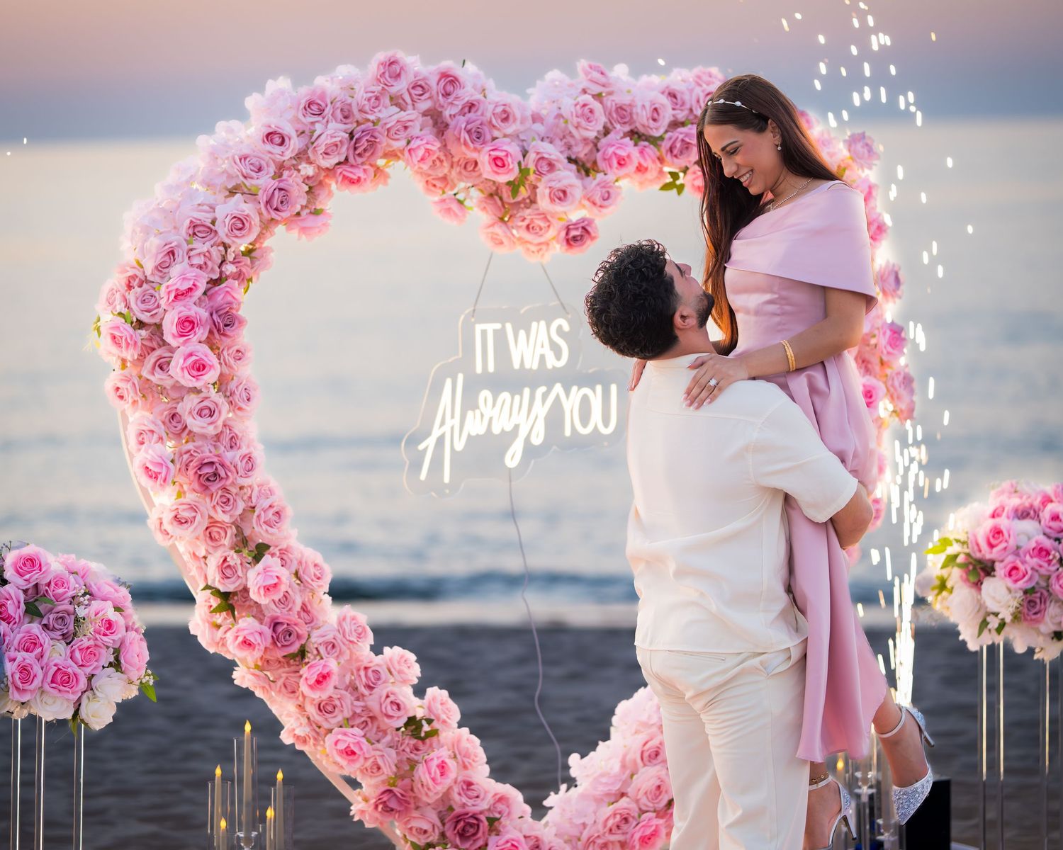 Romantic beach proposal with pink flower arch and sparklers creating a magical sunset atmosphere.