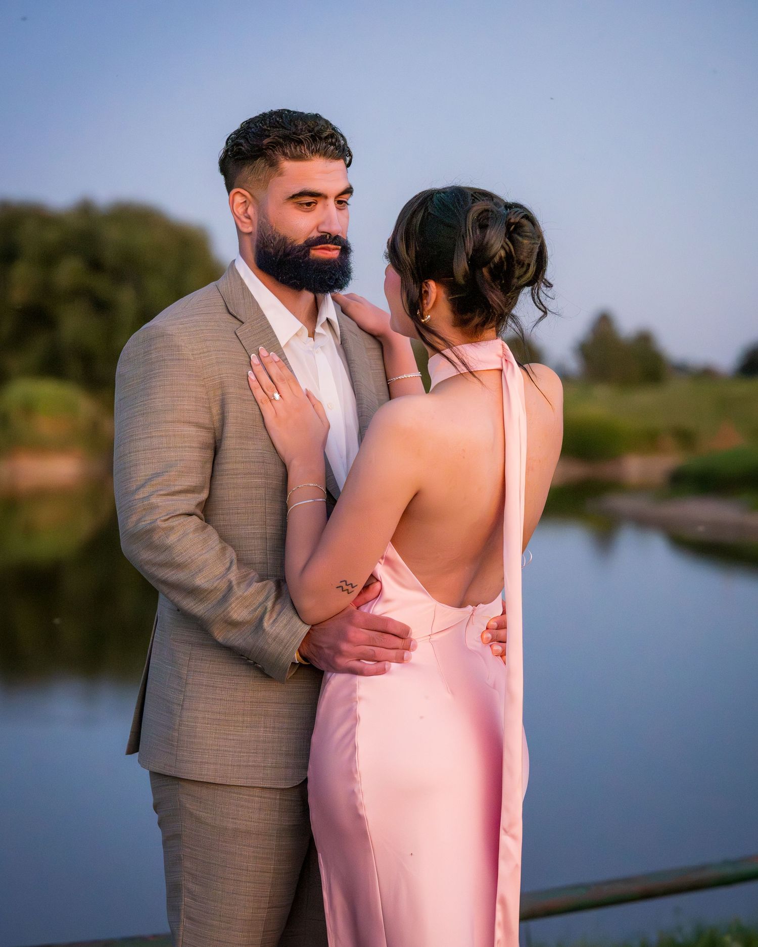 A romantic couple's photoshoot by a lake with one person in a pink dress and another in a tan suit.