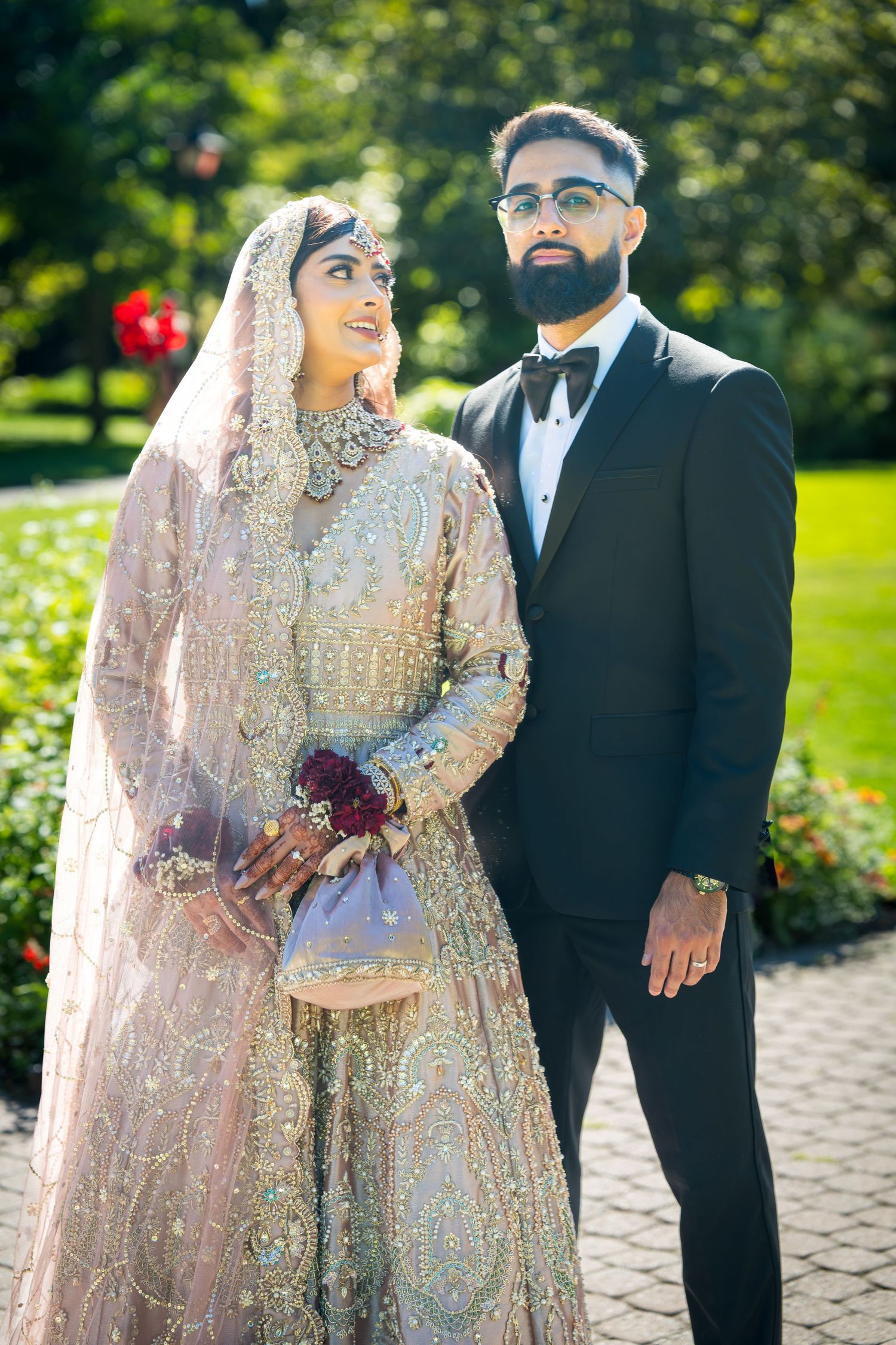 Couple in traditional South Asian wedding attire pose together in an outdoor garden setting.