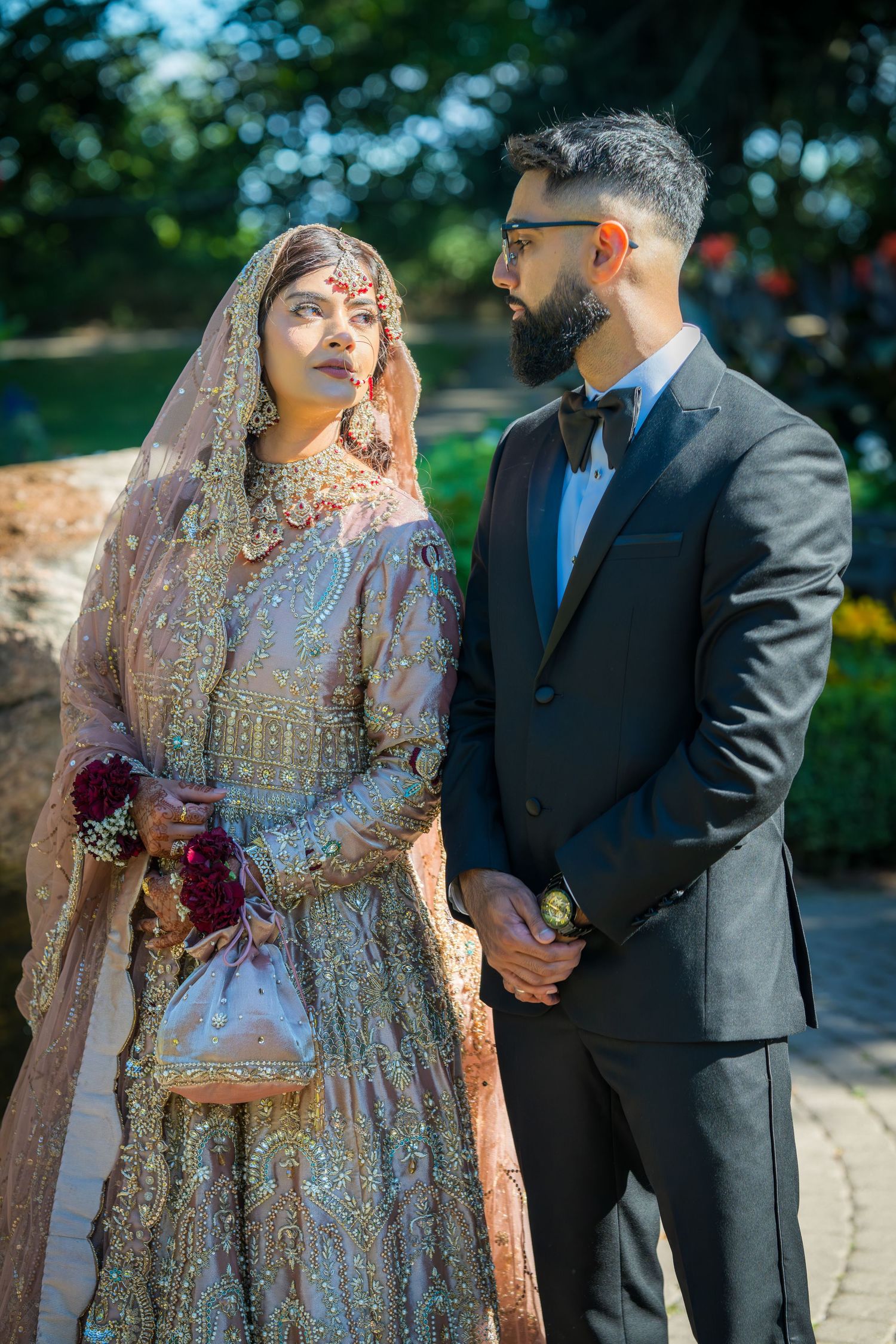 Traditional South Asian wedding couple in elegant attire pose outdoors with bride in ornate lehenga and groom in suit.