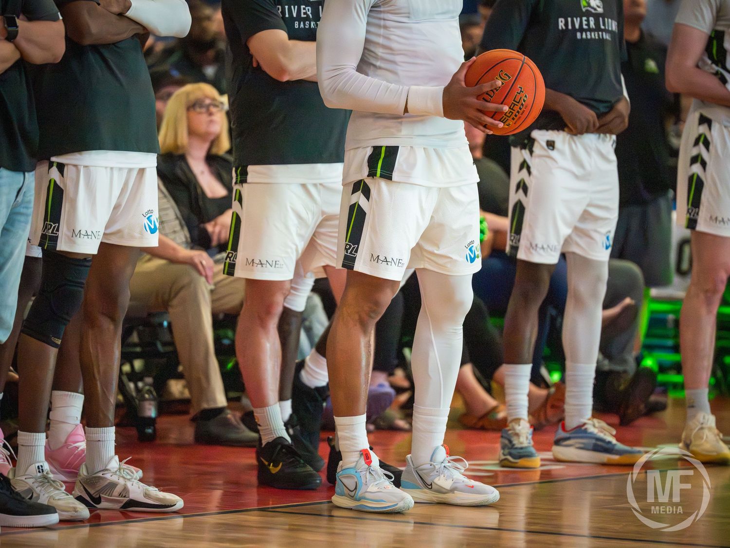 River Lions basketball players stand on the sideline in white and green uniforms during a CEBL game.