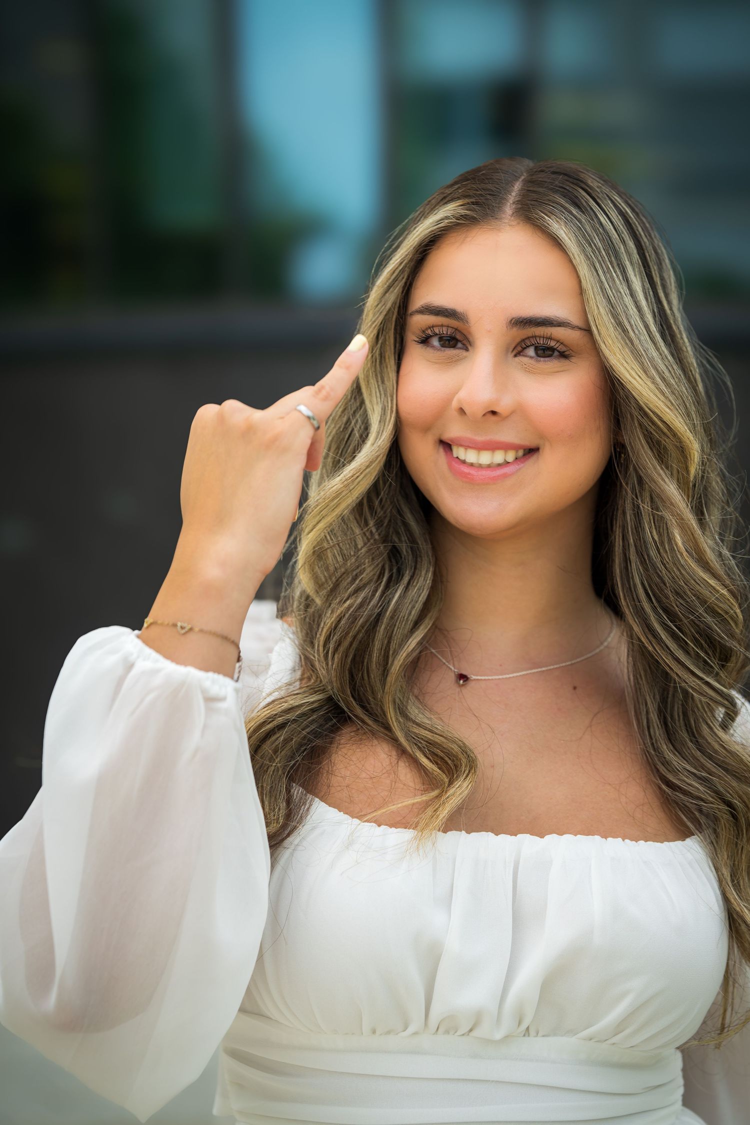 Graduate in white dress with flowing sleeves poses against modern architectural background.