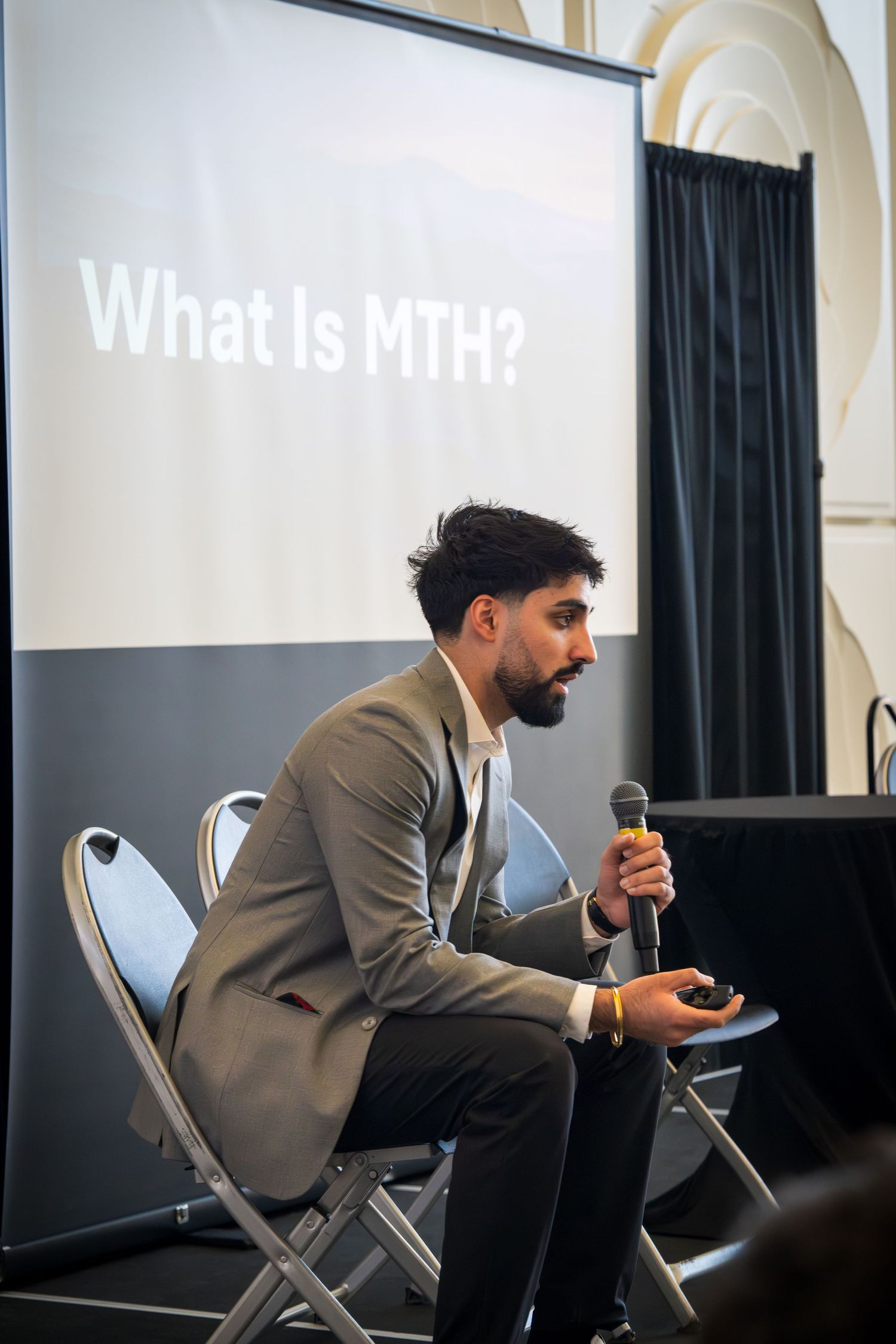 Business presenter sitting on stage with projection screen showing 'What is MFN?' text.