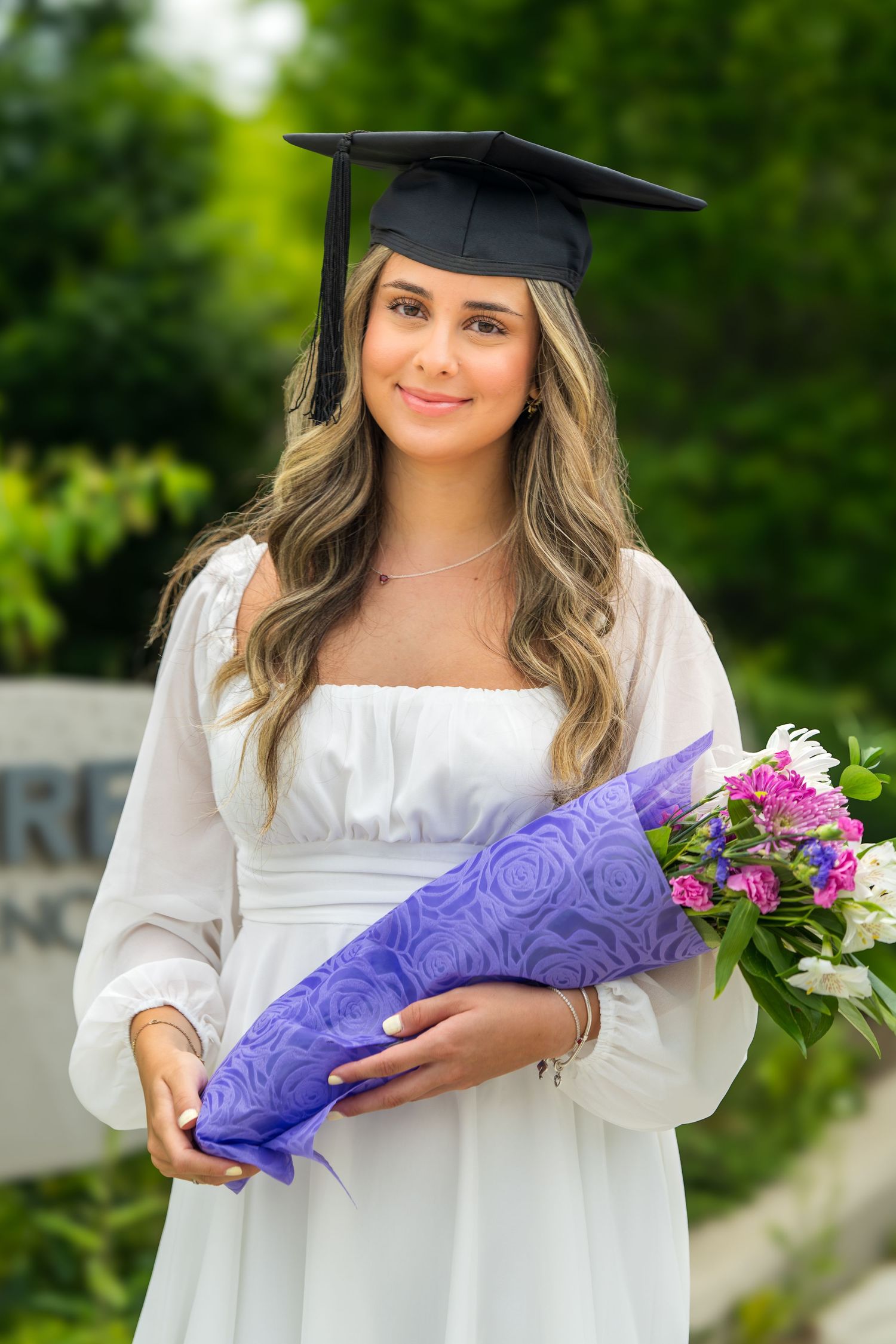 Graduate in white dress holds purple bouquet wrap and flowers while wearing cap and gown.