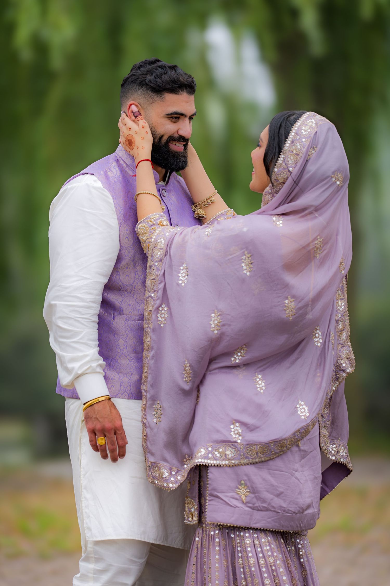 A couple in matching lavender and white traditional dress share an affectionate moment under willow trees.