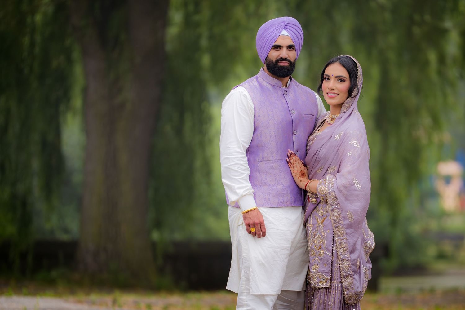 A couple shares a tender moment in matching purple and white traditional Indian clothing under willow trees.