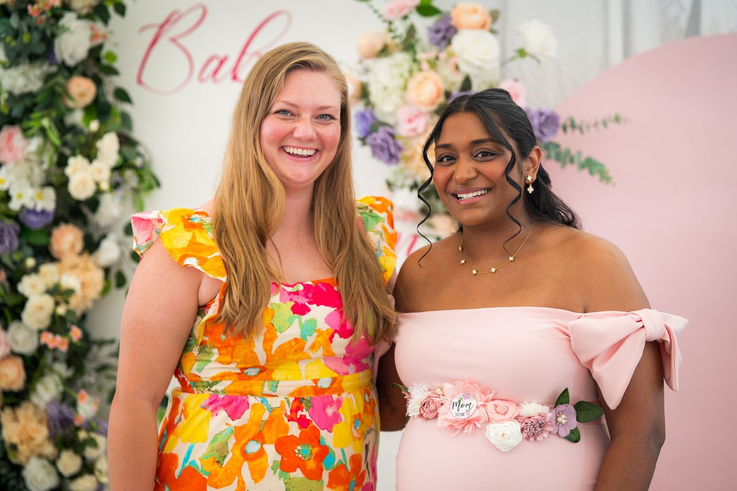 Guests in colorful dresses pose together at a baby shower with floral decorations.