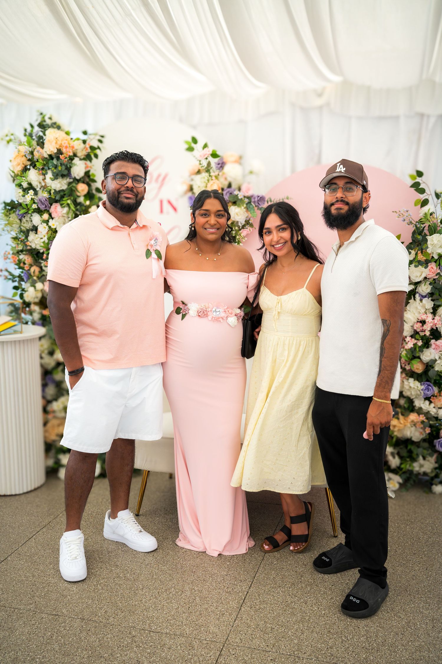 Four guests pose at baby celebration with elegant floral arch and white draping.
