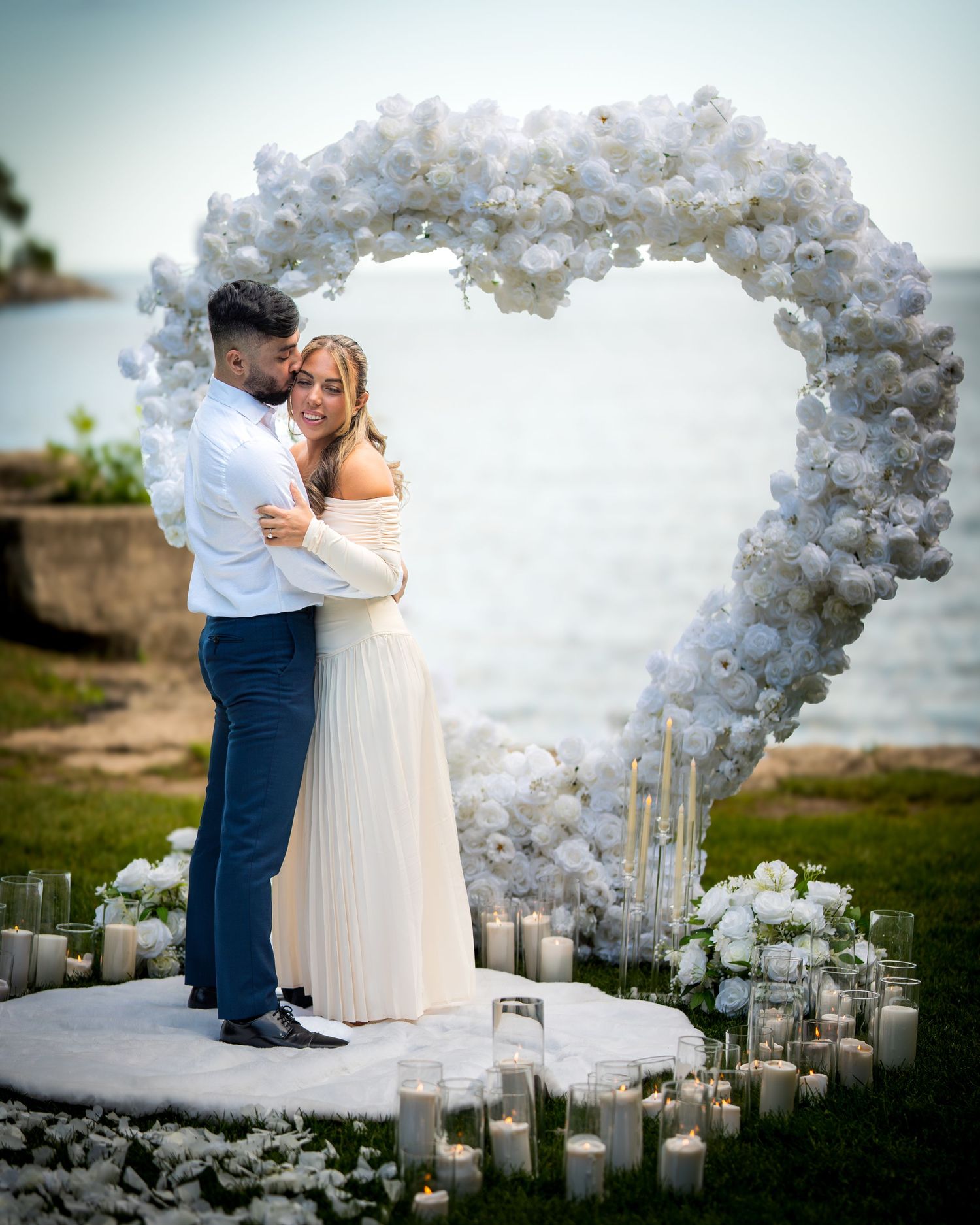 Romantic proposal scene with white balloon heart arch and glowing candles by the ocean.