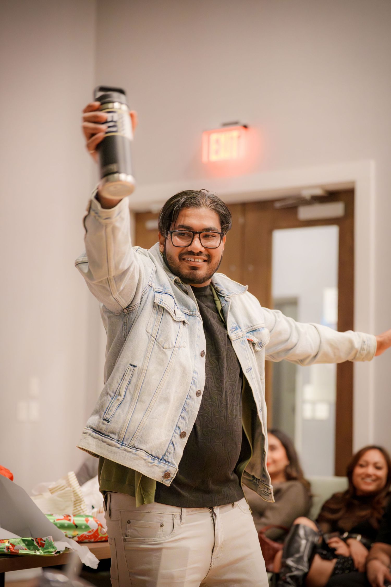 Person in denim jacket celebrating with raised arms at an indoor party.