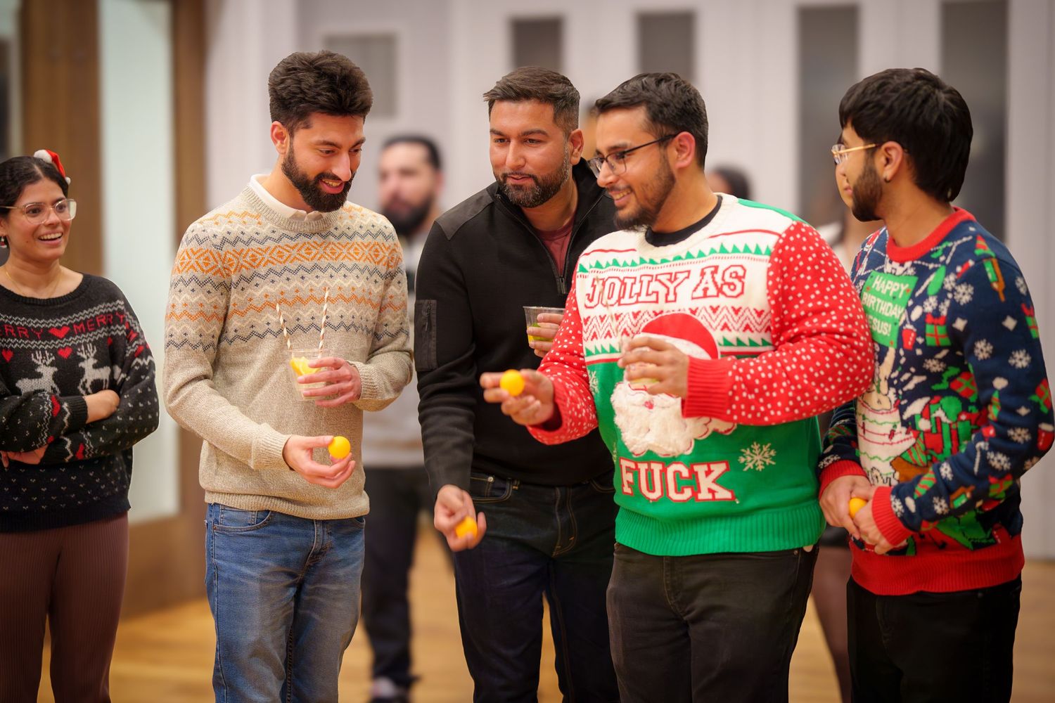 People wearing colorful holiday sweaters gather at indoor social event.